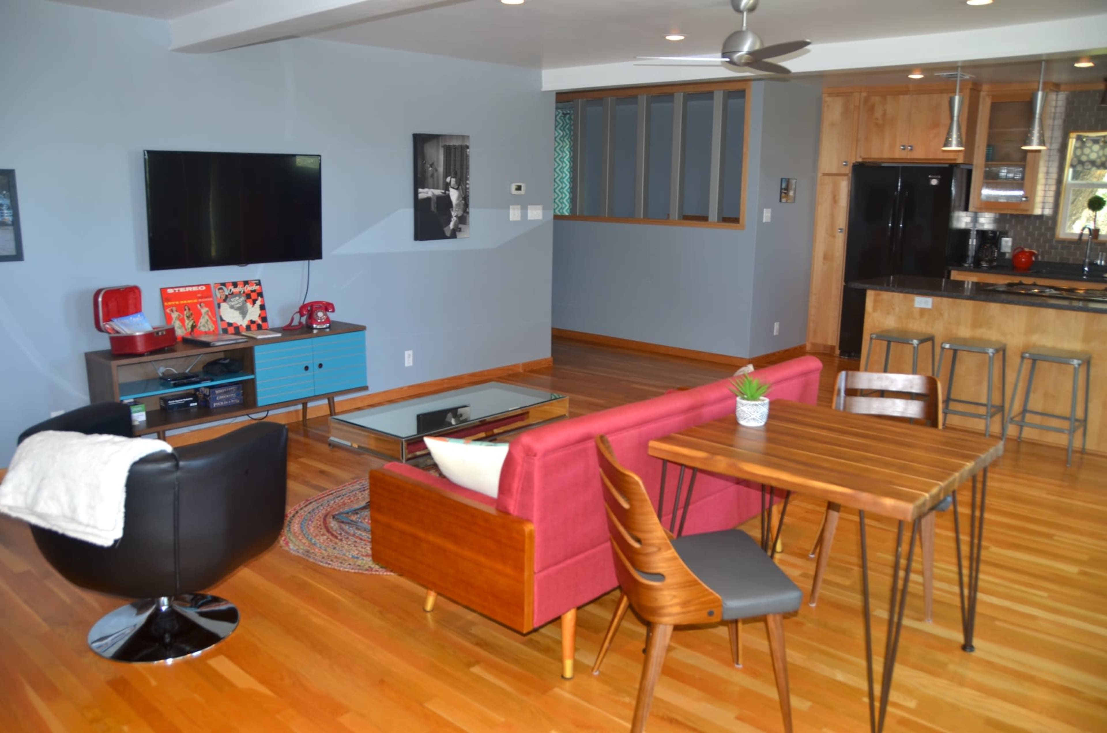 The interior of a modern living area features a television, a dining table, a sofa, and a kitchen with a bar counter, all arranged on hardwood floors.