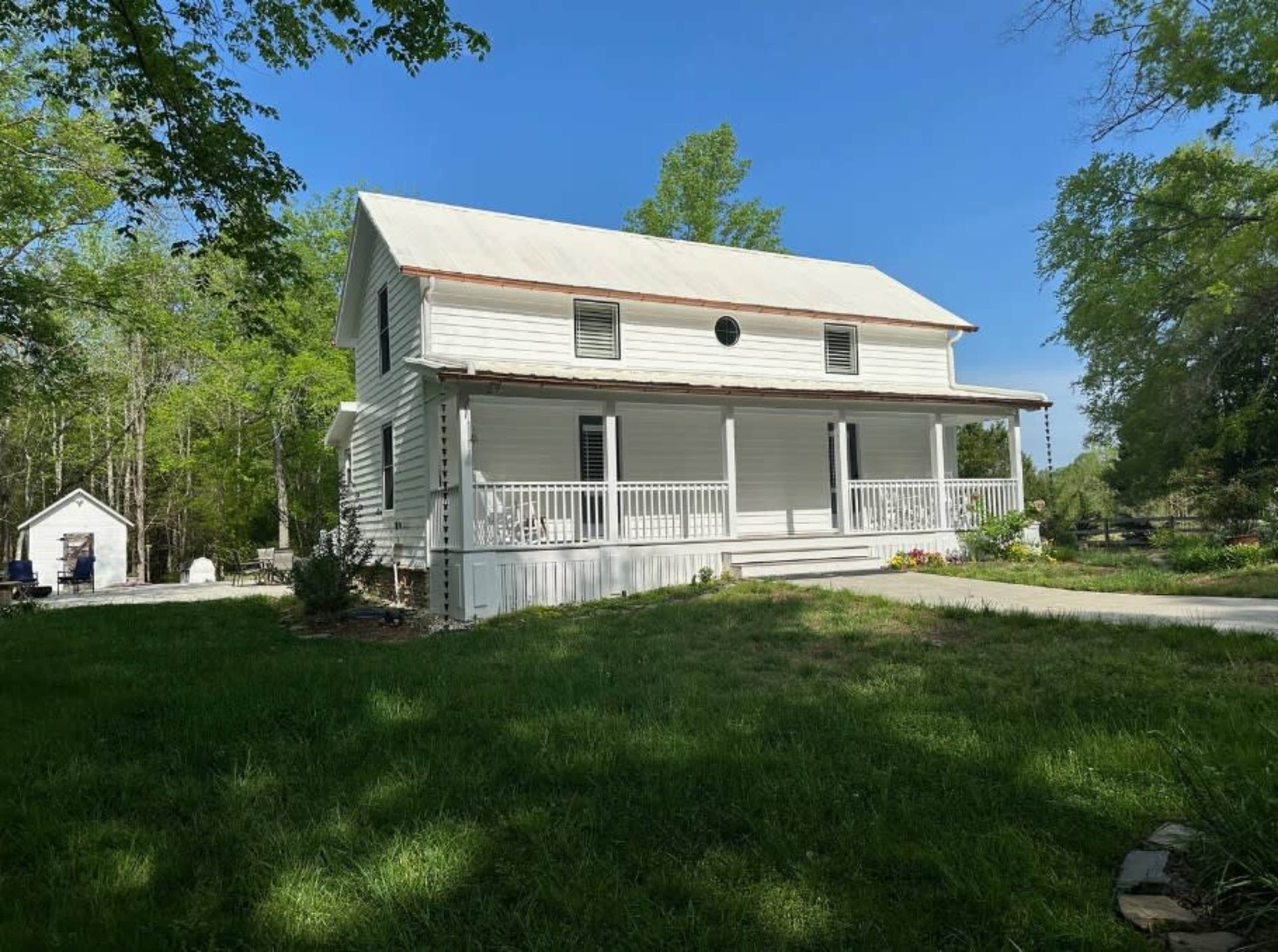 A two-story white house with a front porch and a metal roof stands on a grassy lawn surrounded by trees.