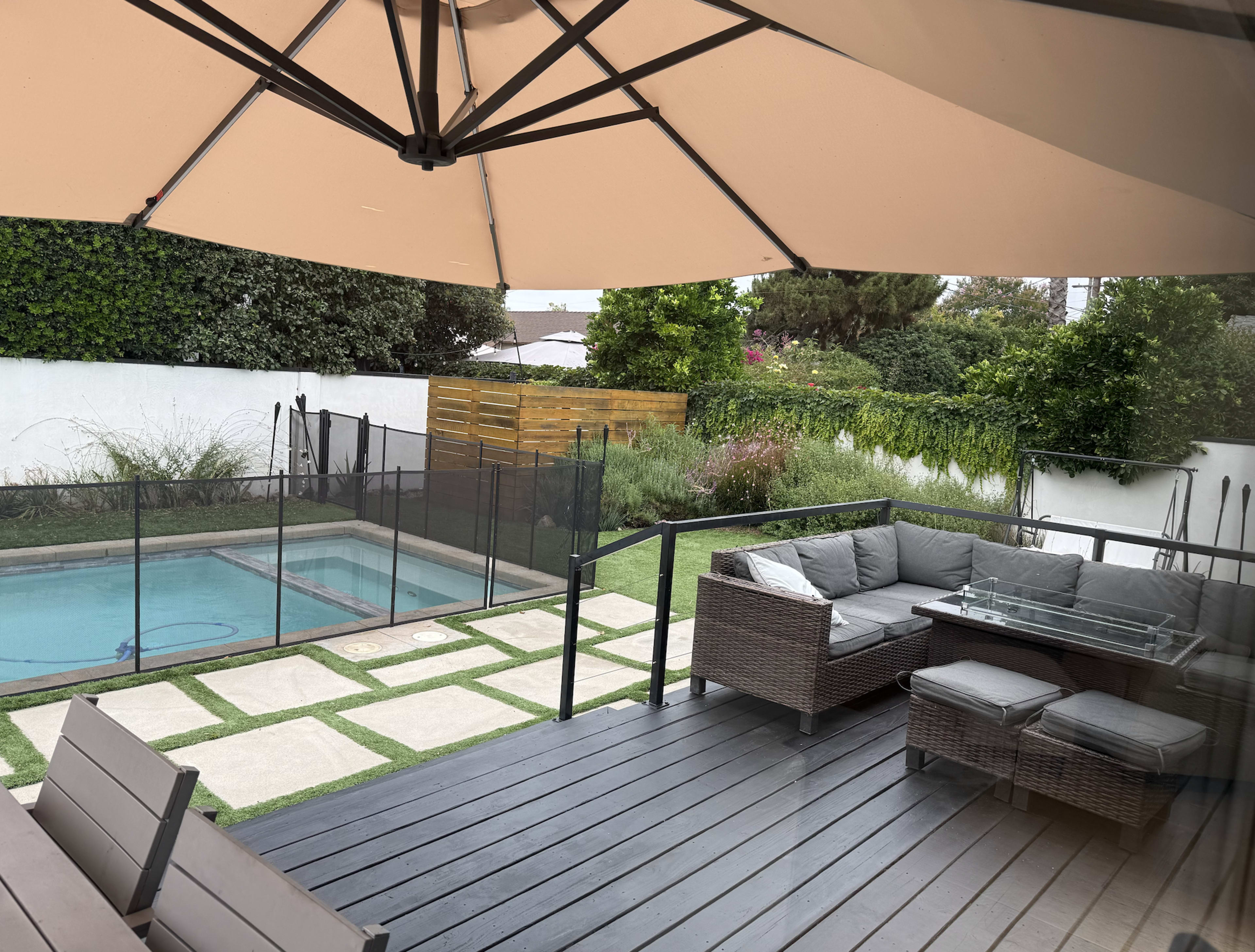 A patio features a seating area with a gray wicker sofa and glass table overlooking a swimming pool surrounded by a safety fence and landscaped garden.