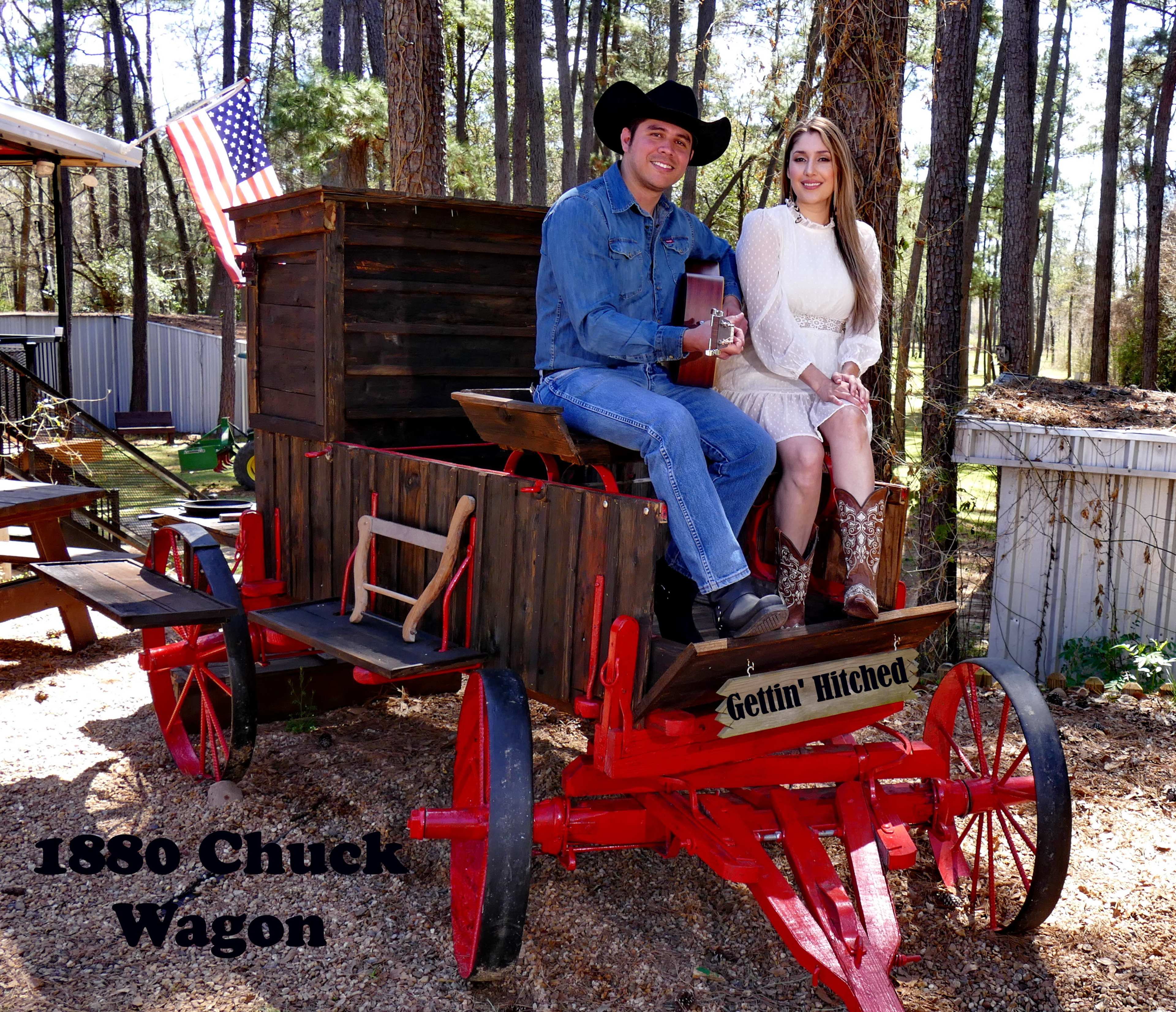 A man and a woman sit together on an antique red chuck wagon in a wooded area, with an American flag in the background.