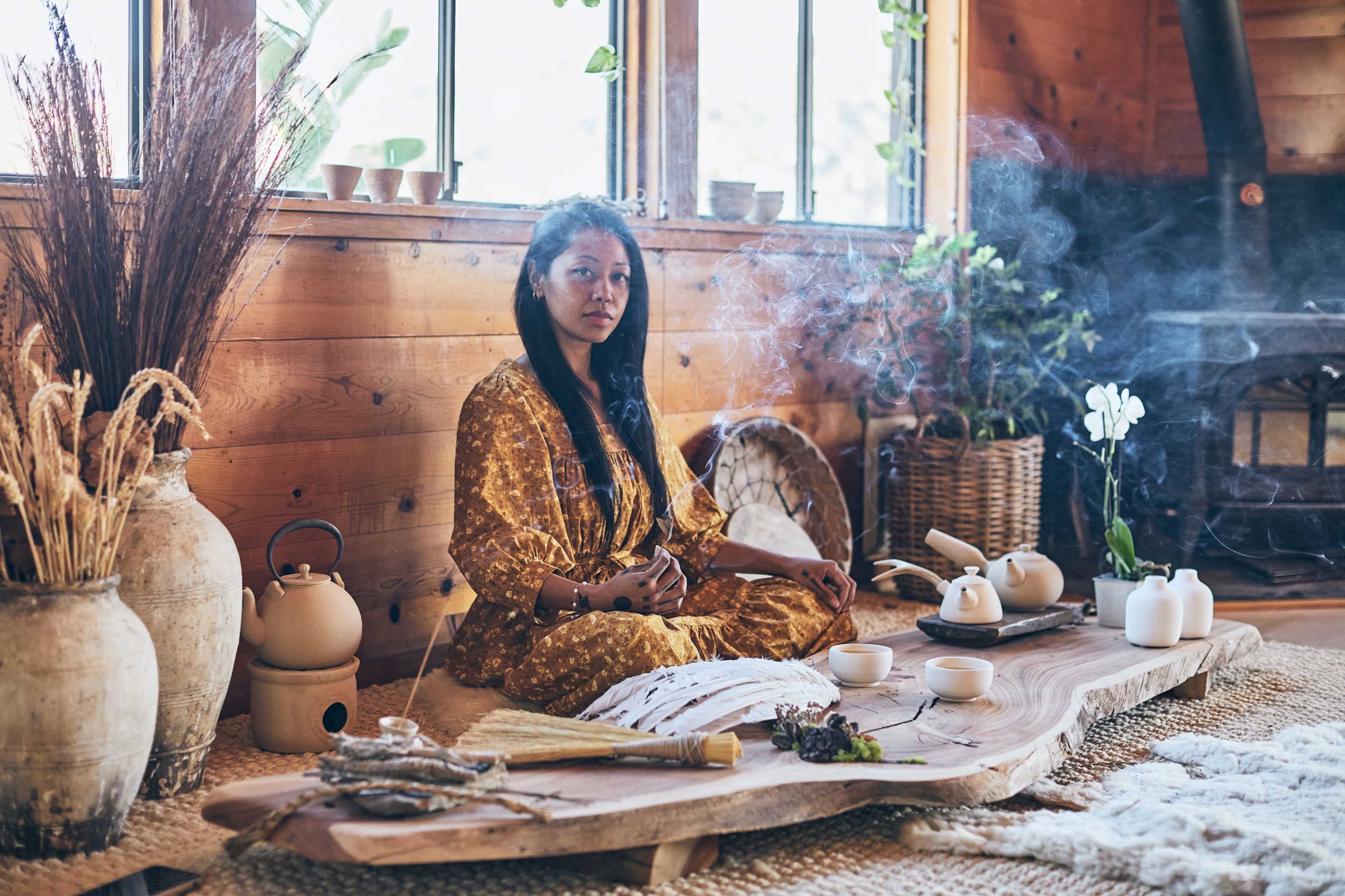 A woman sits cross-legged on a wooden floor in a warmly lit room surrounded by plants, while tea is being prepared on a large wooden table with smoke rising from incense.