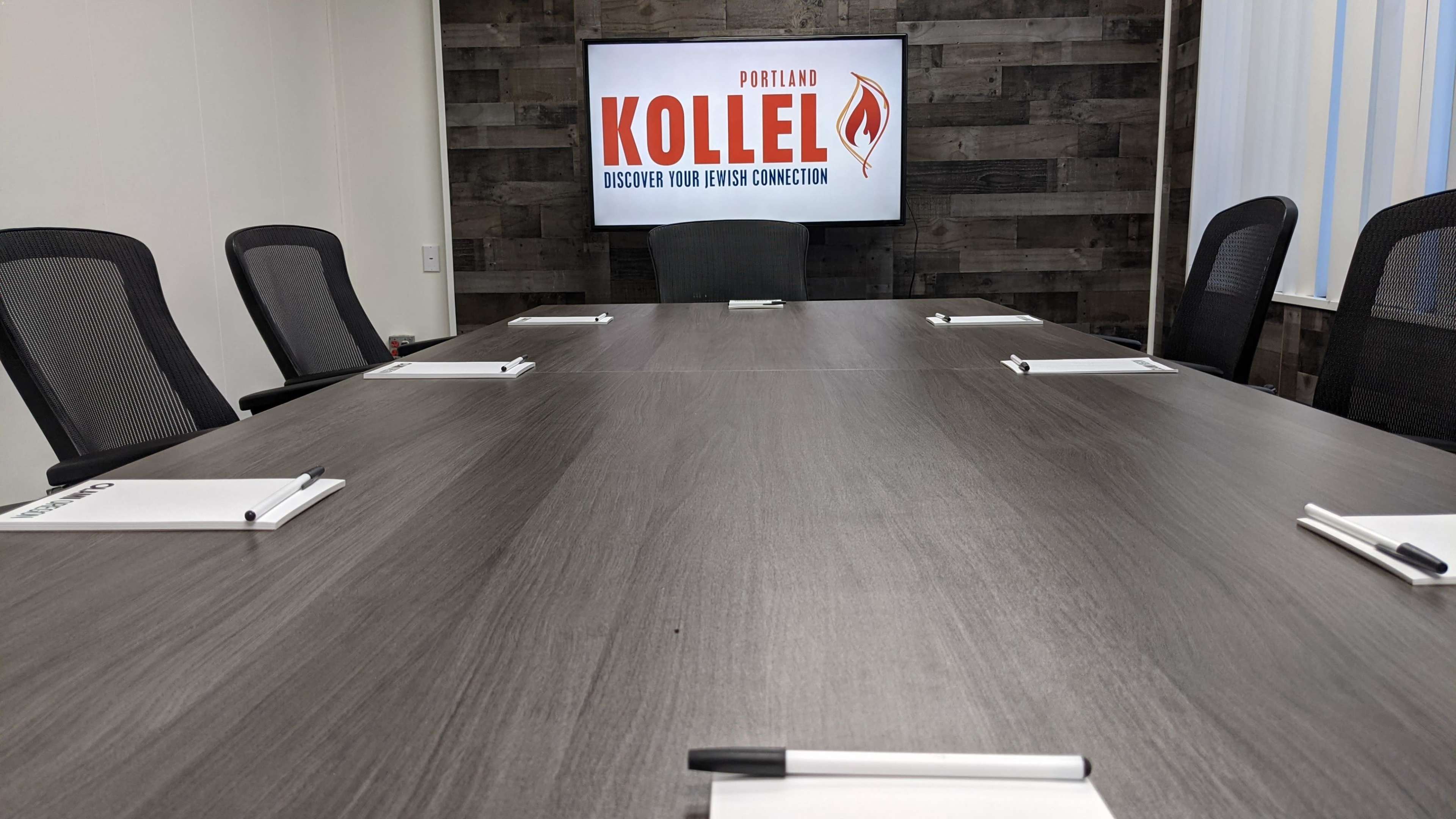 A conference room with a large table, black chairs, and a screen displaying "Portland Kollel" on the wall.
