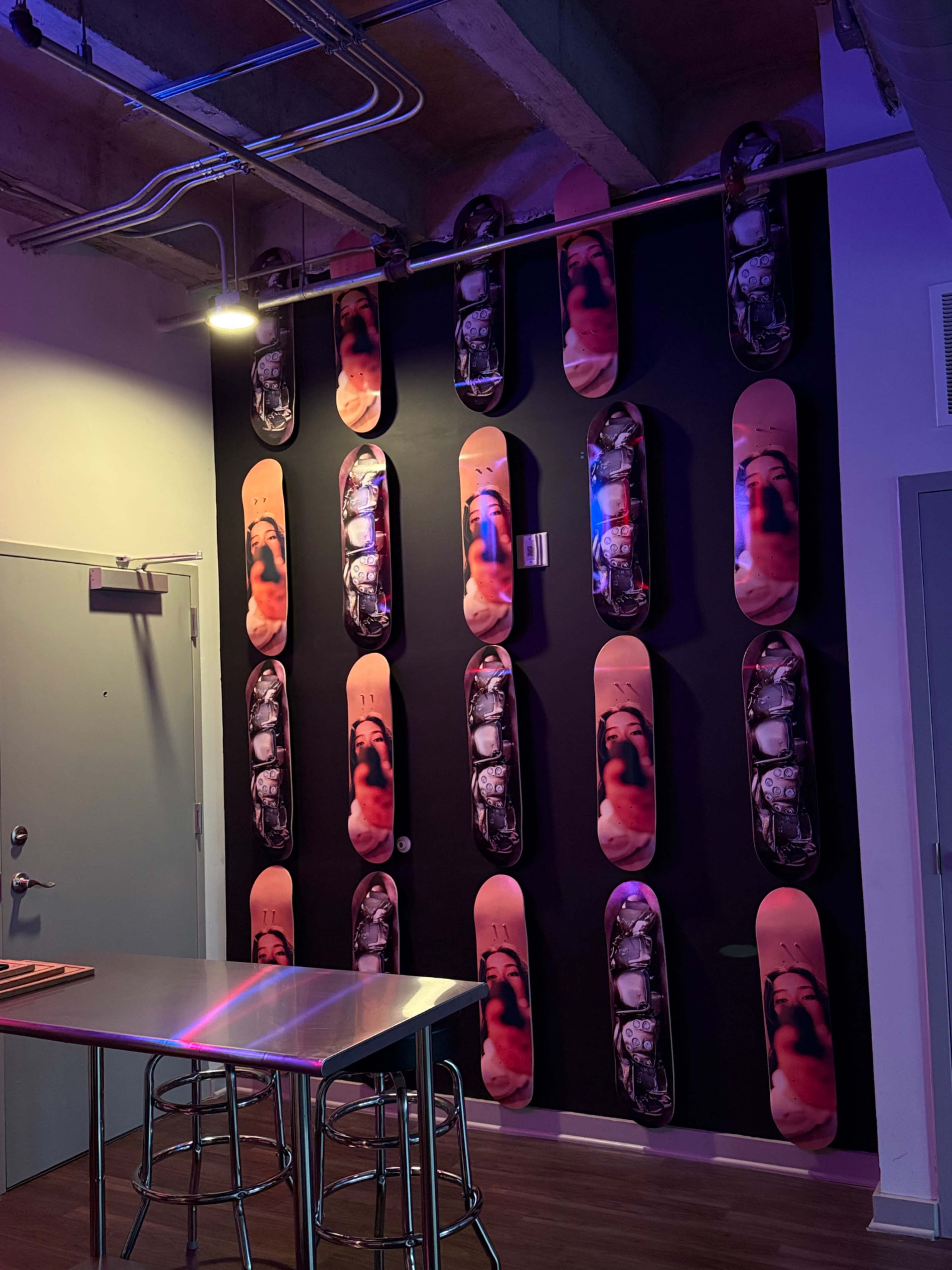 The image shows a wall decorated with various skateboard decks featuring artistic designs, arranged in a grid pattern, with a nearby metal table and stools.