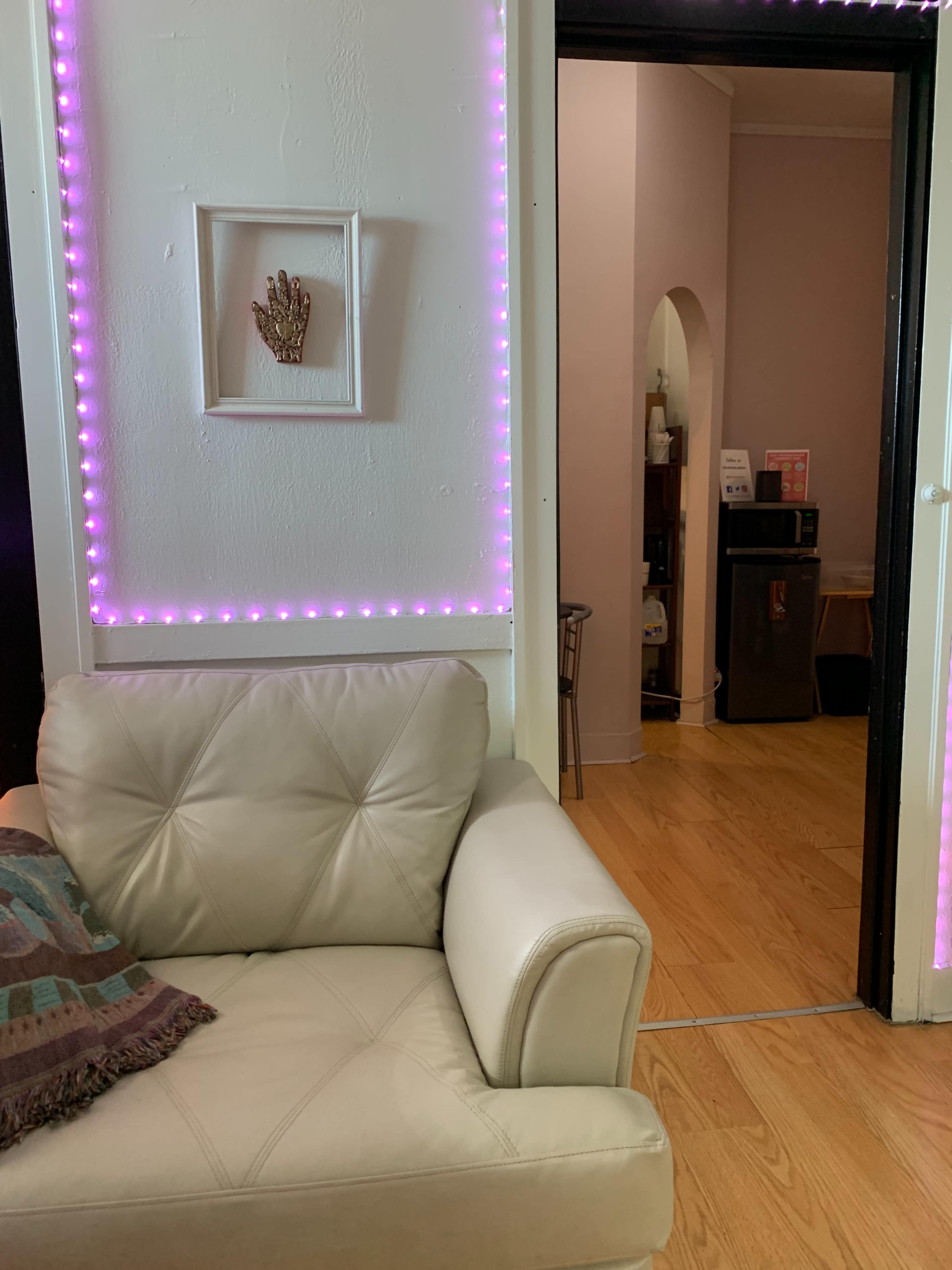 A white leather sofa with a decorative throw pillow is positioned near a doorway framed by pink LED lights, leading to an adjacent room with a small refrigerator.