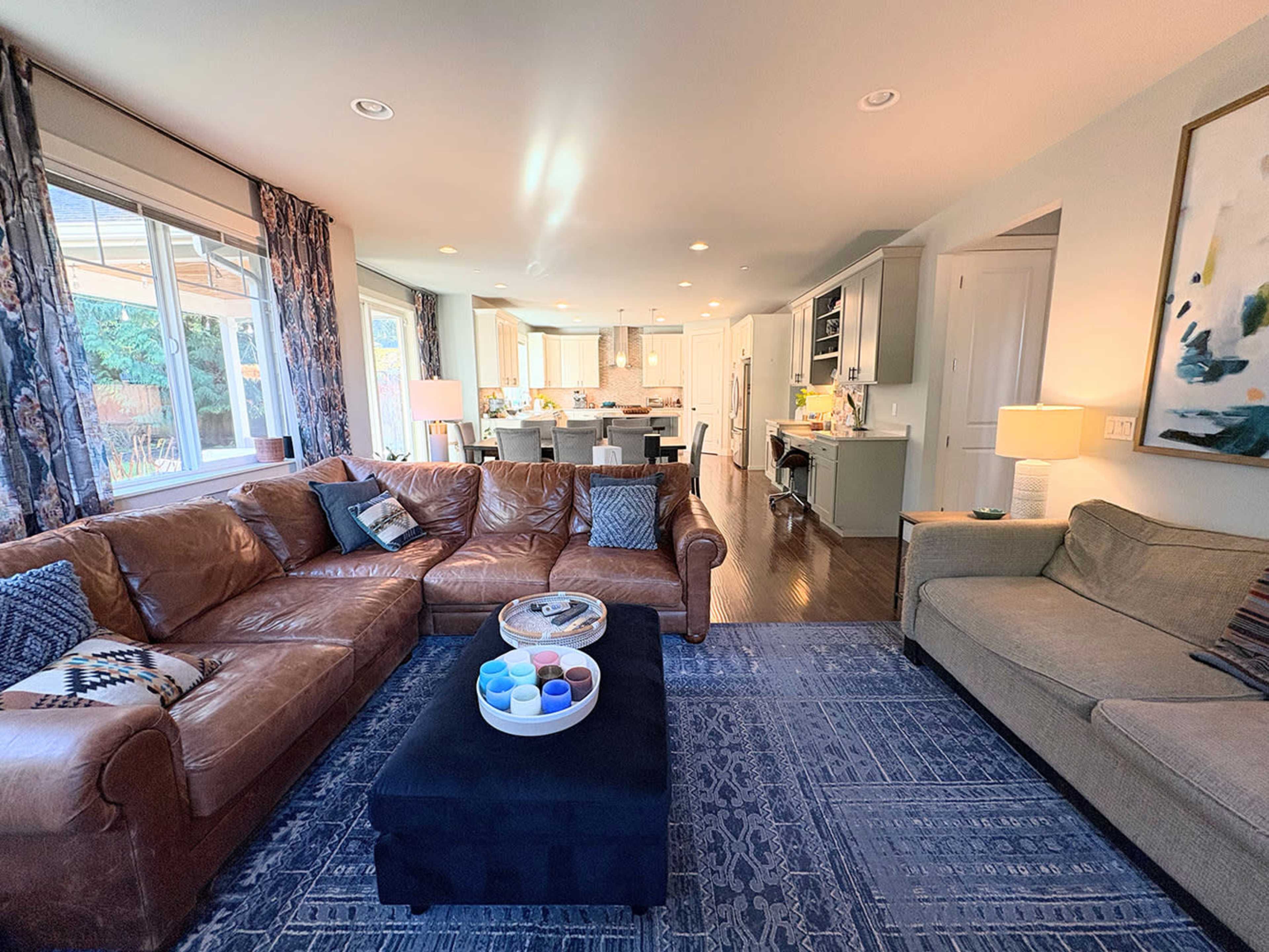A spacious living room features a brown leather sectional sofa, a black ottoman, and a view into a modern kitchen with light cabinetry and a dining area.