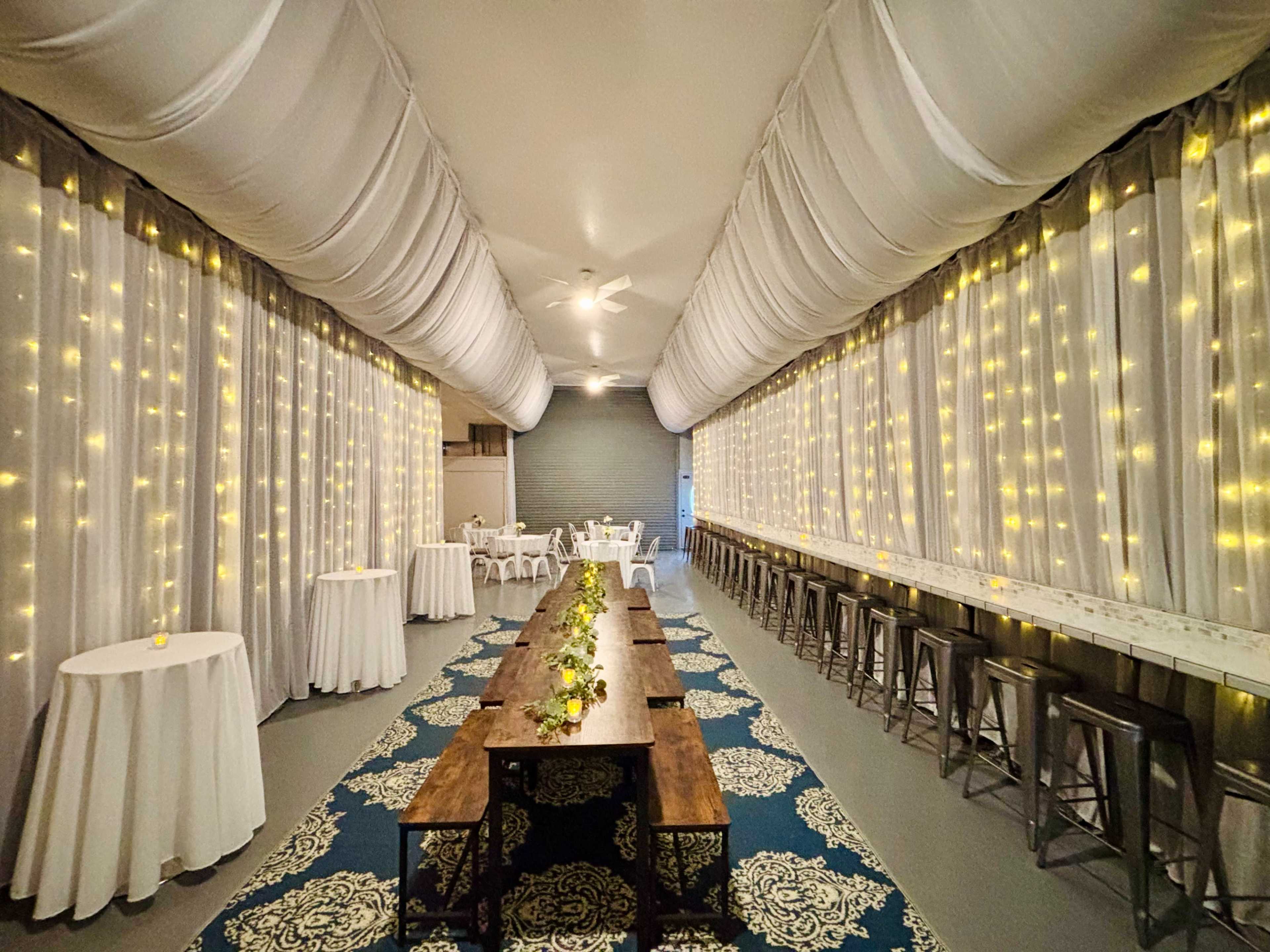 The image shows a beautifully decorated indoor event space with draped fabric, string lights, and tables arranged for dining.