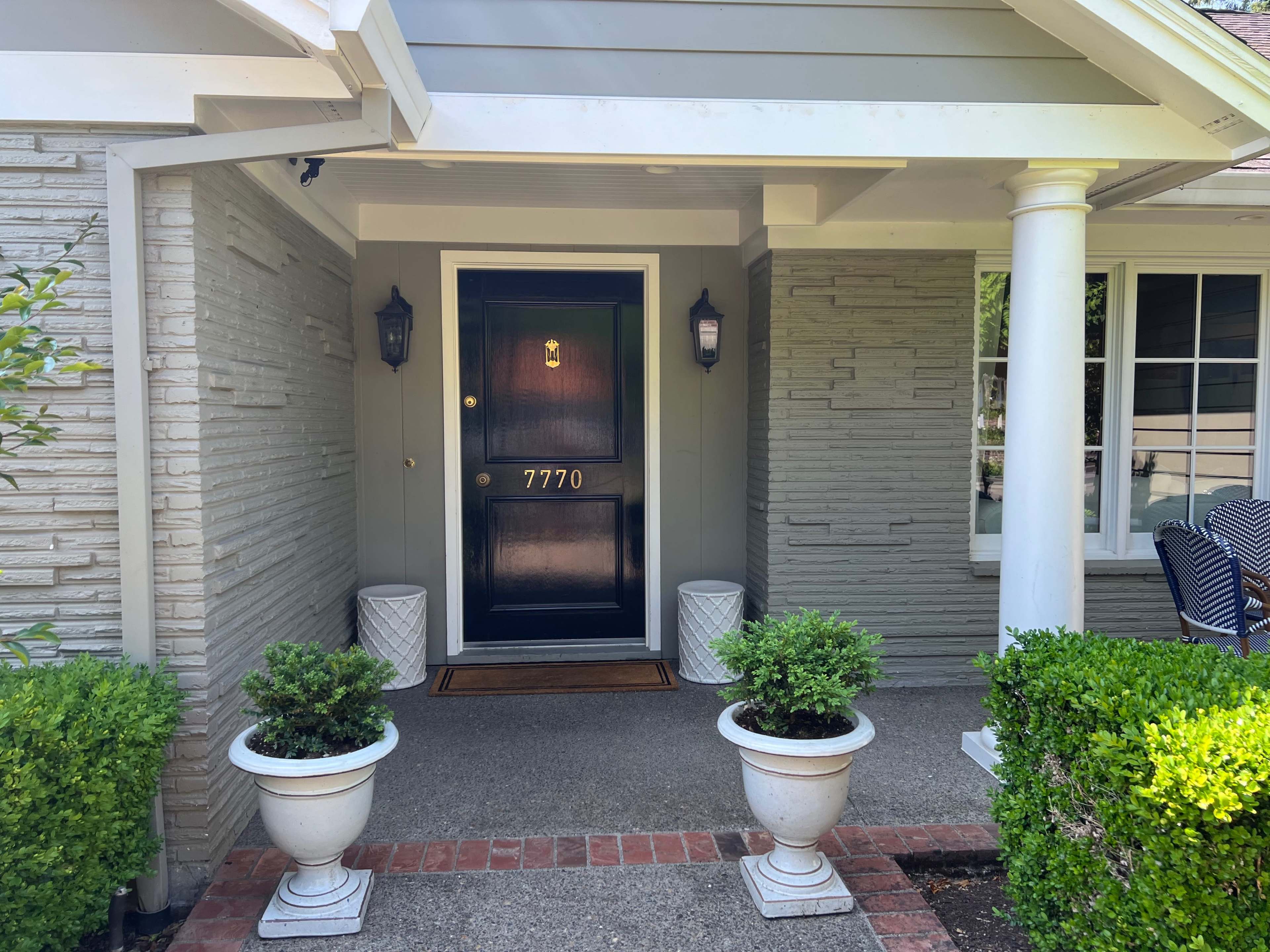 A house entrance with a dark door marked "7770," flanked by two potted shrubs and decorative light fixtures.