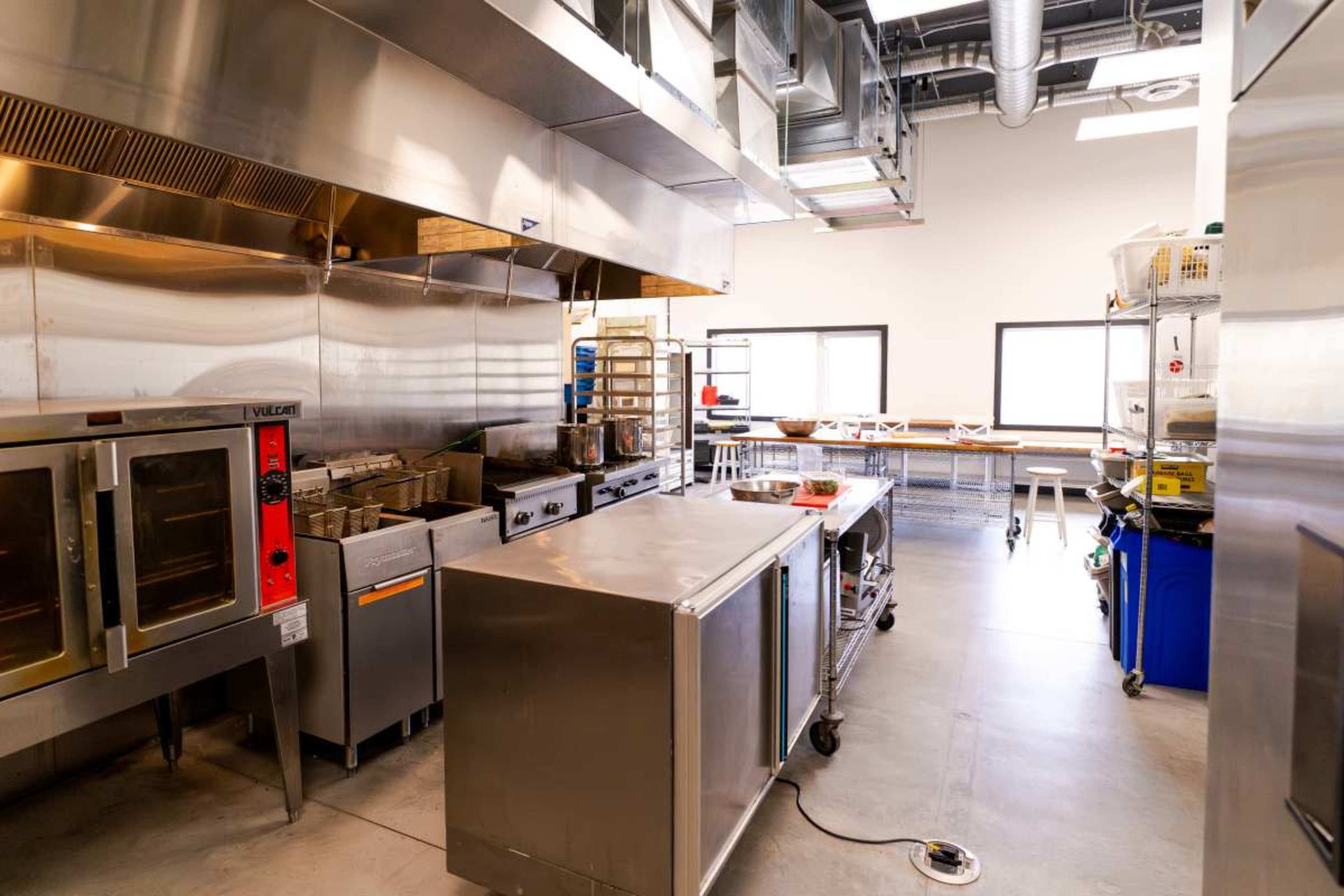 The image shows a commercial kitchen with stainless steel appliances, including ovens and a preparation area, illuminated by natural light from large windows.