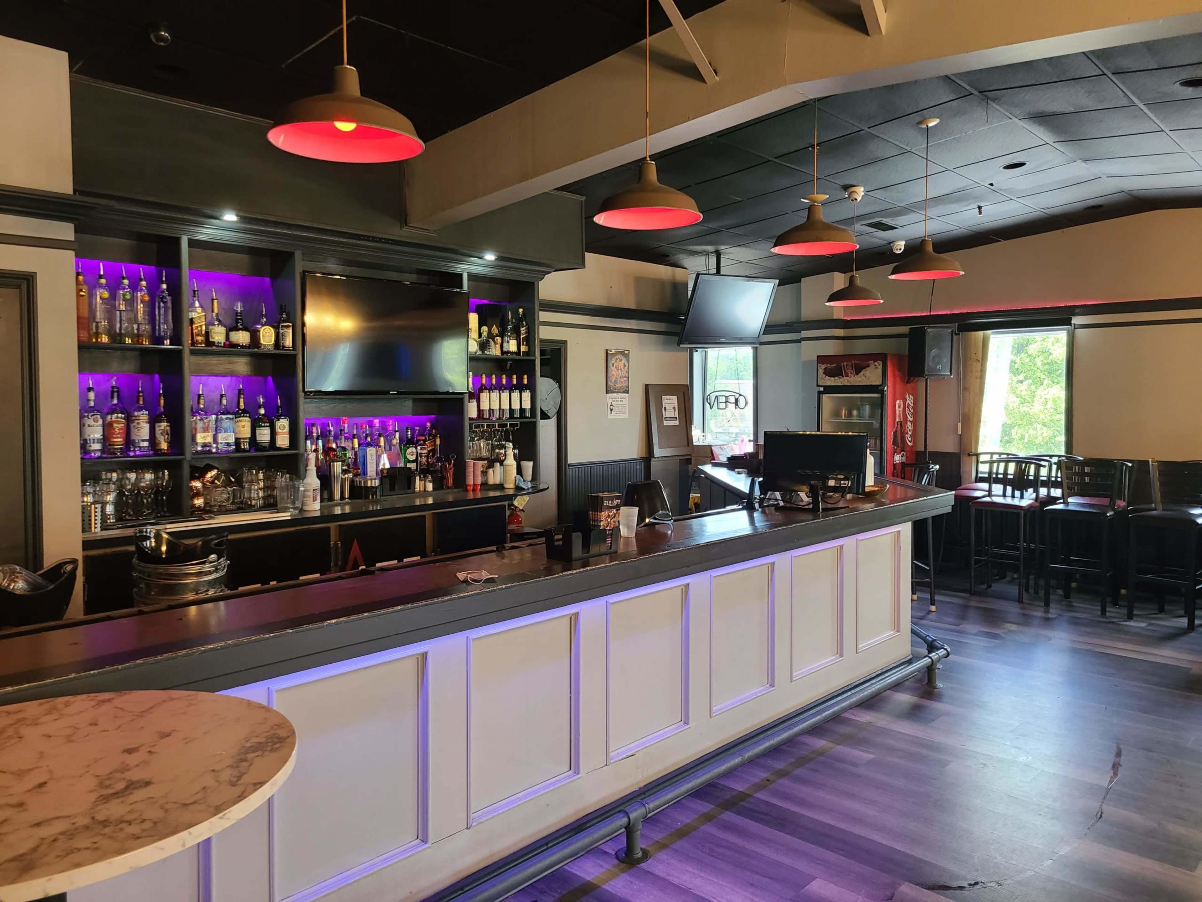 A well-lit bar area with a long countertop, shelves stocked with various liquor bottles, and several seating areas in the background.