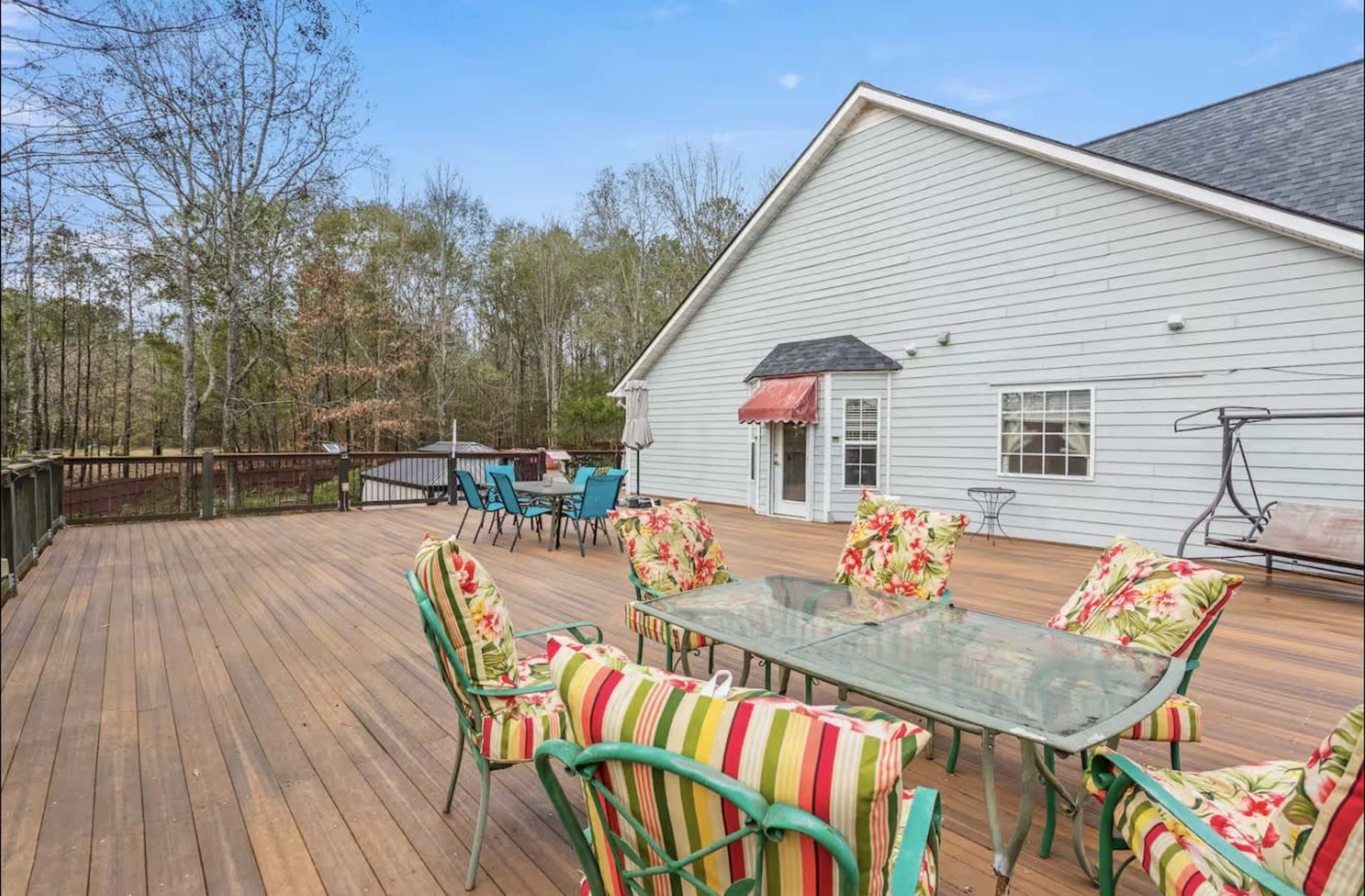 A spacious wooden deck features a large glass table surrounded by colorful chairs, with a house and wooded area in the background.