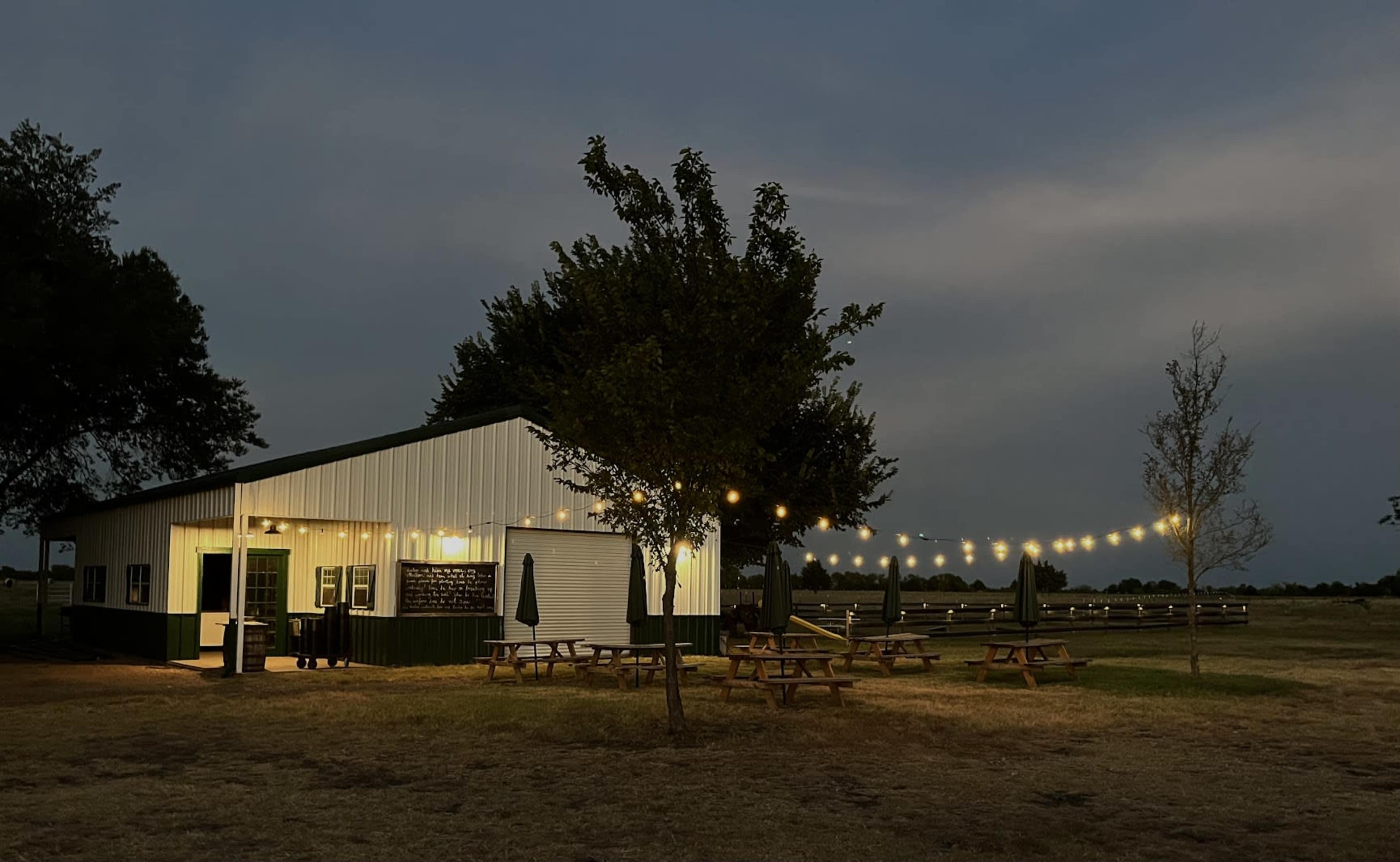 A small, well-lit building with string lights outside is surrounded by picnic tables in a grassy area at dusk.