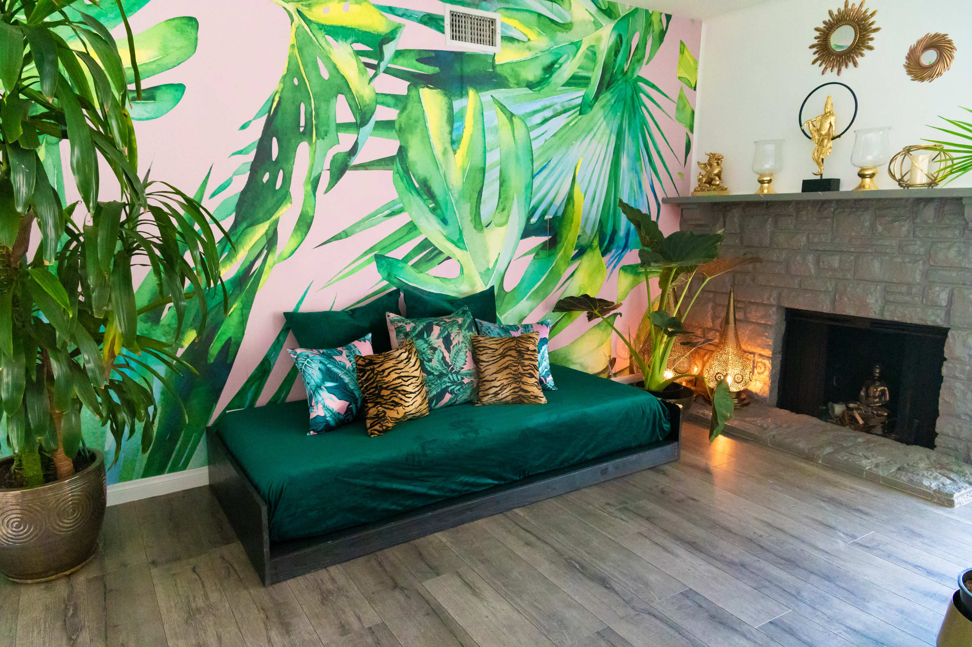 A living space with a green daybed adorned with decorative pillows in front of a stone fireplace, surrounded by large plants and a vibrant mural of tropical leaves on the wall.