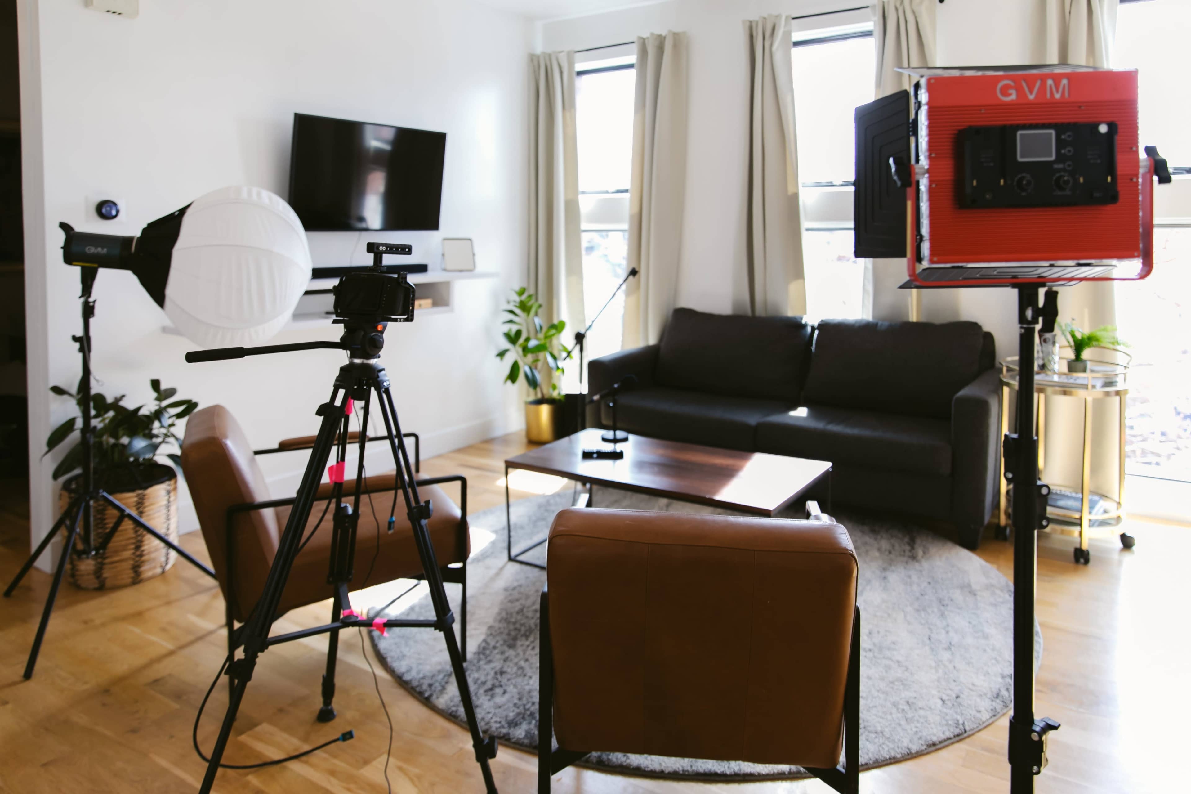A living room is set up for filming, featuring a camera on a tripod, lighting equipment, and a sofa surrounded by plants.
