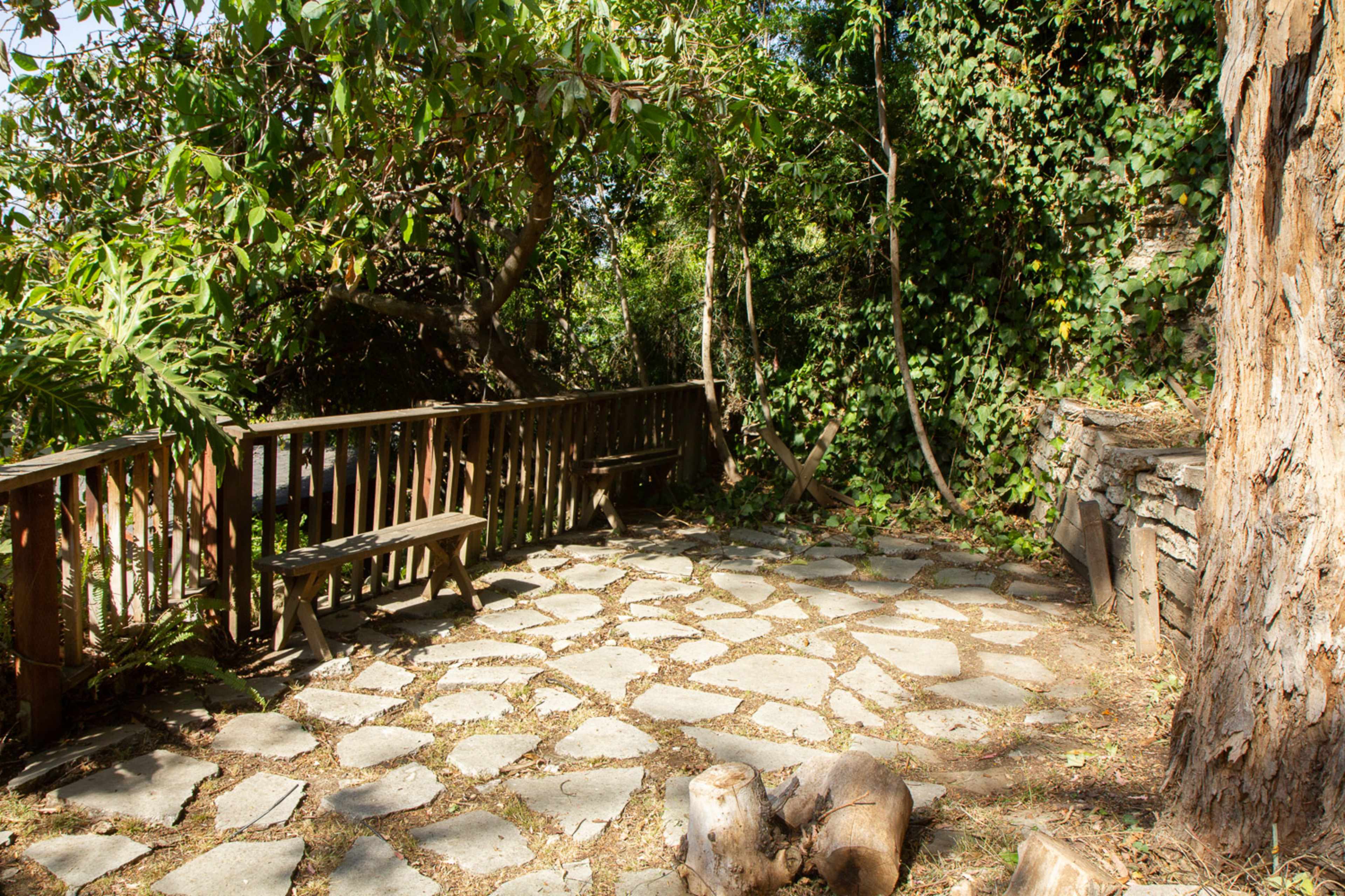 A stone patio surrounded by greenery, with wooden benches positioned along the railing and a tree on one side.