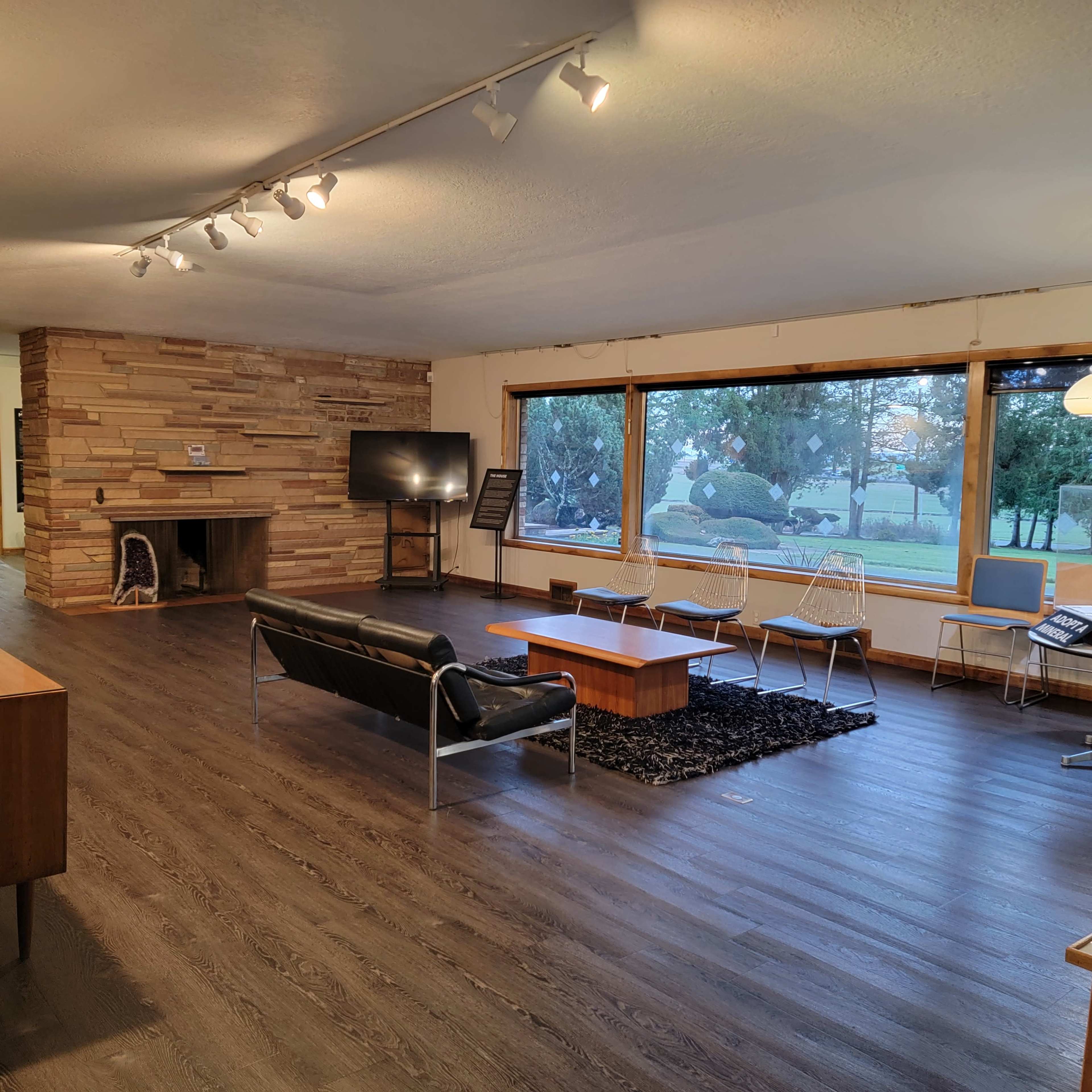 The image shows a modern living room with a stone fireplace, large windows overlooking a garden, and minimalist furniture including a black sofa and several chairs.
