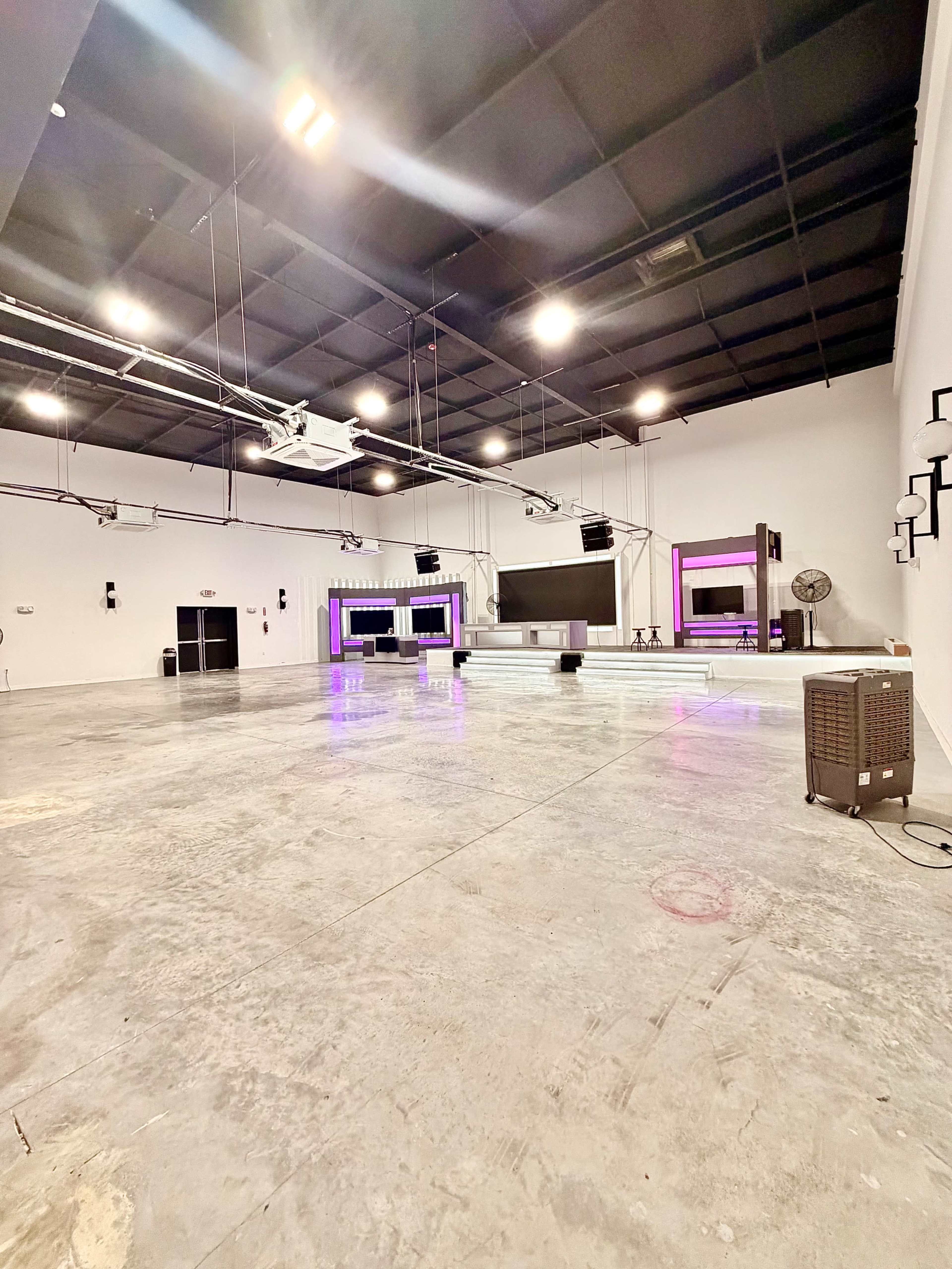 The image shows an empty indoor space with a concrete floor, featuring a stage area with large screens and lights at the front.