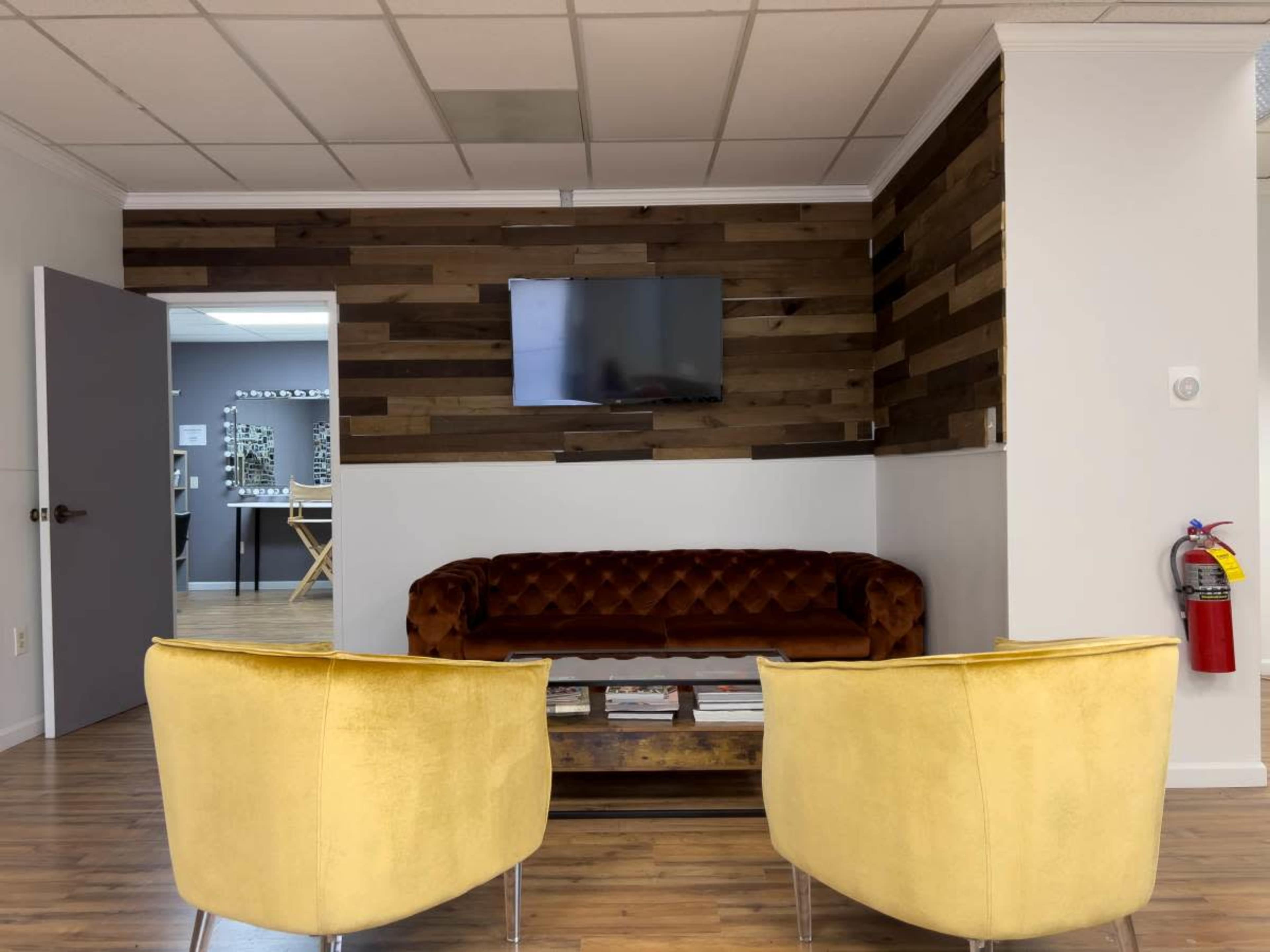 The image shows a waiting area with a dark red couch and two yellow chairs facing a wall-mounted television and a wooden accent wall.
