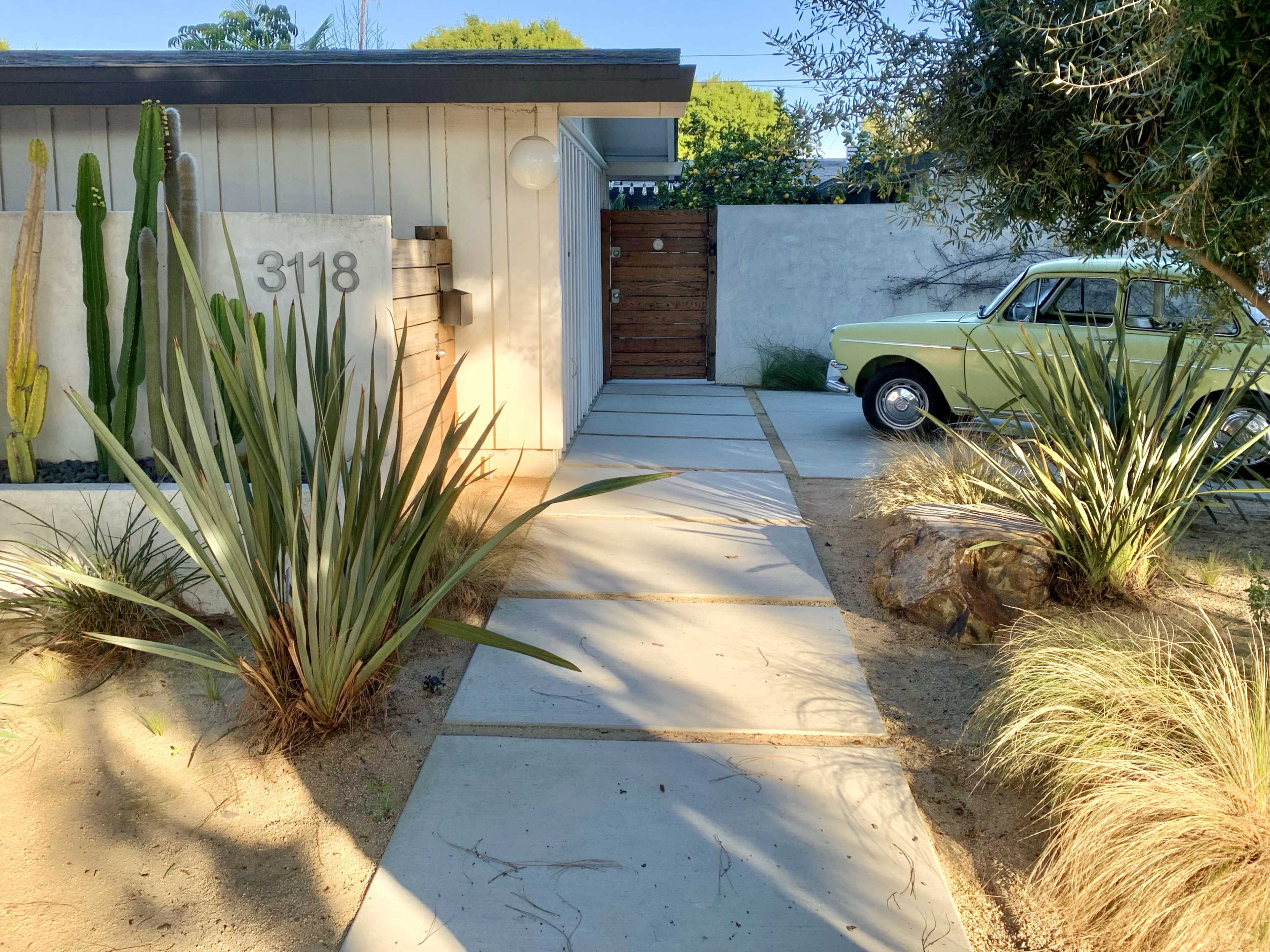 A concrete walkway leads to a modern house with a wooden door and a vintage car parked nearby, surrounded by desert plants and gravel landscaping.