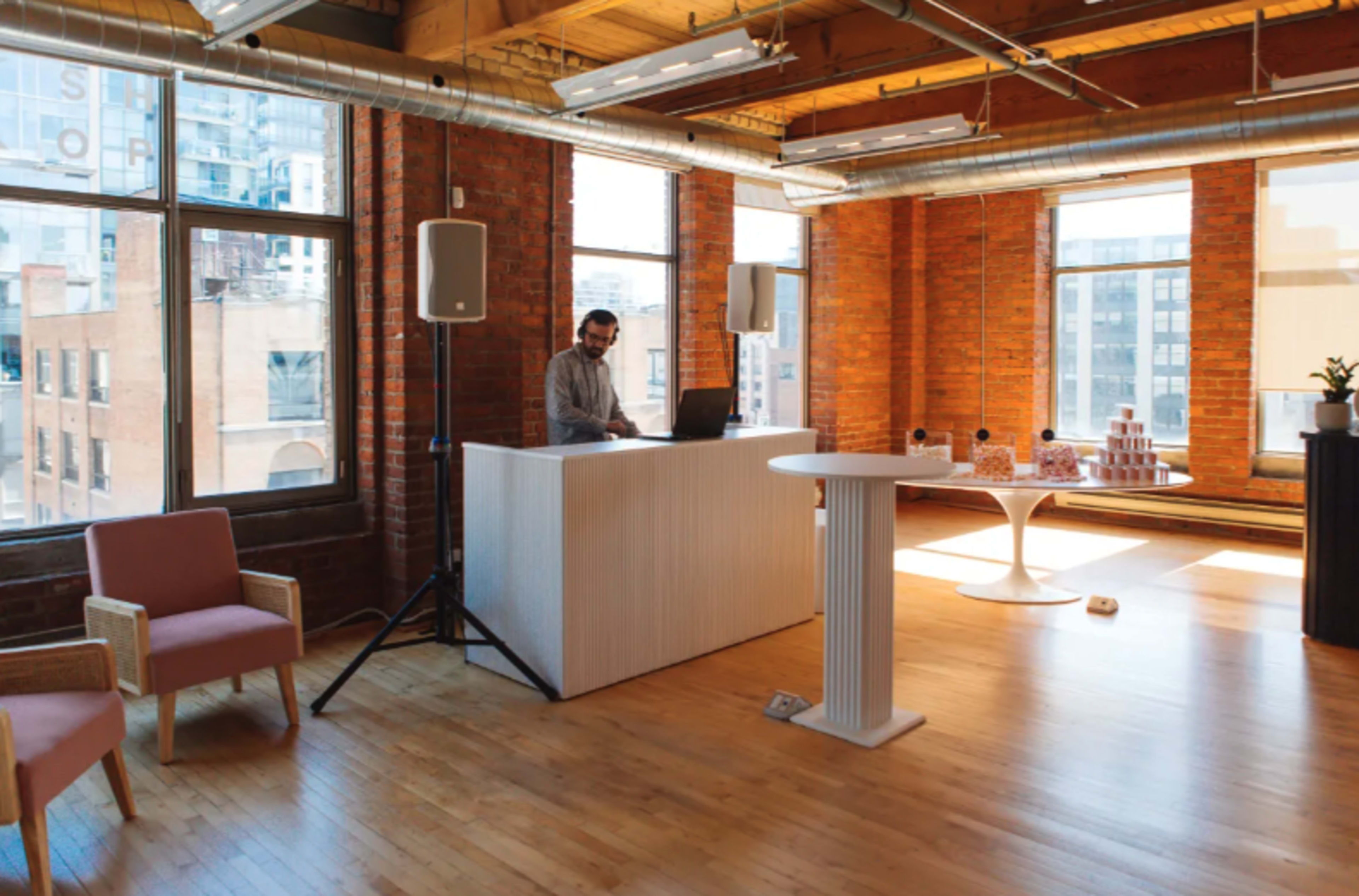 A man stands behind a white reception desk in a spacious, well-lit room with large windows and wooden floors.