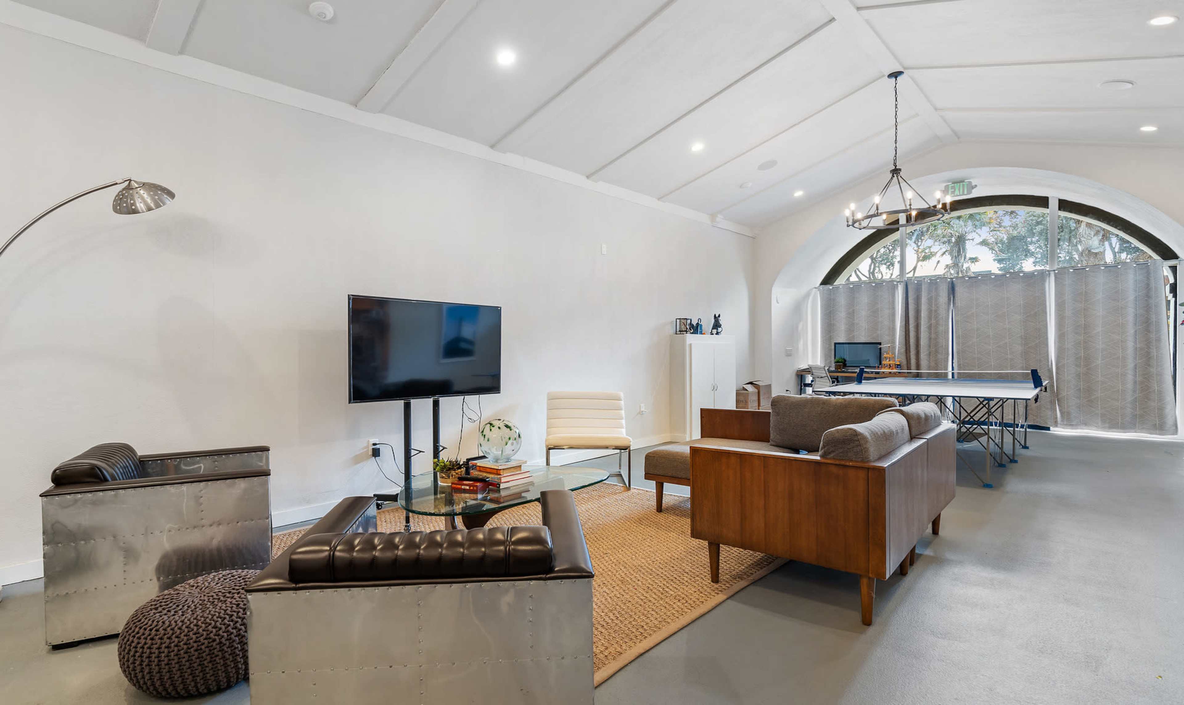 The scene features a modern living space with a television, seating area, and a ping pong table, illuminated by natural light through a large arched window.
