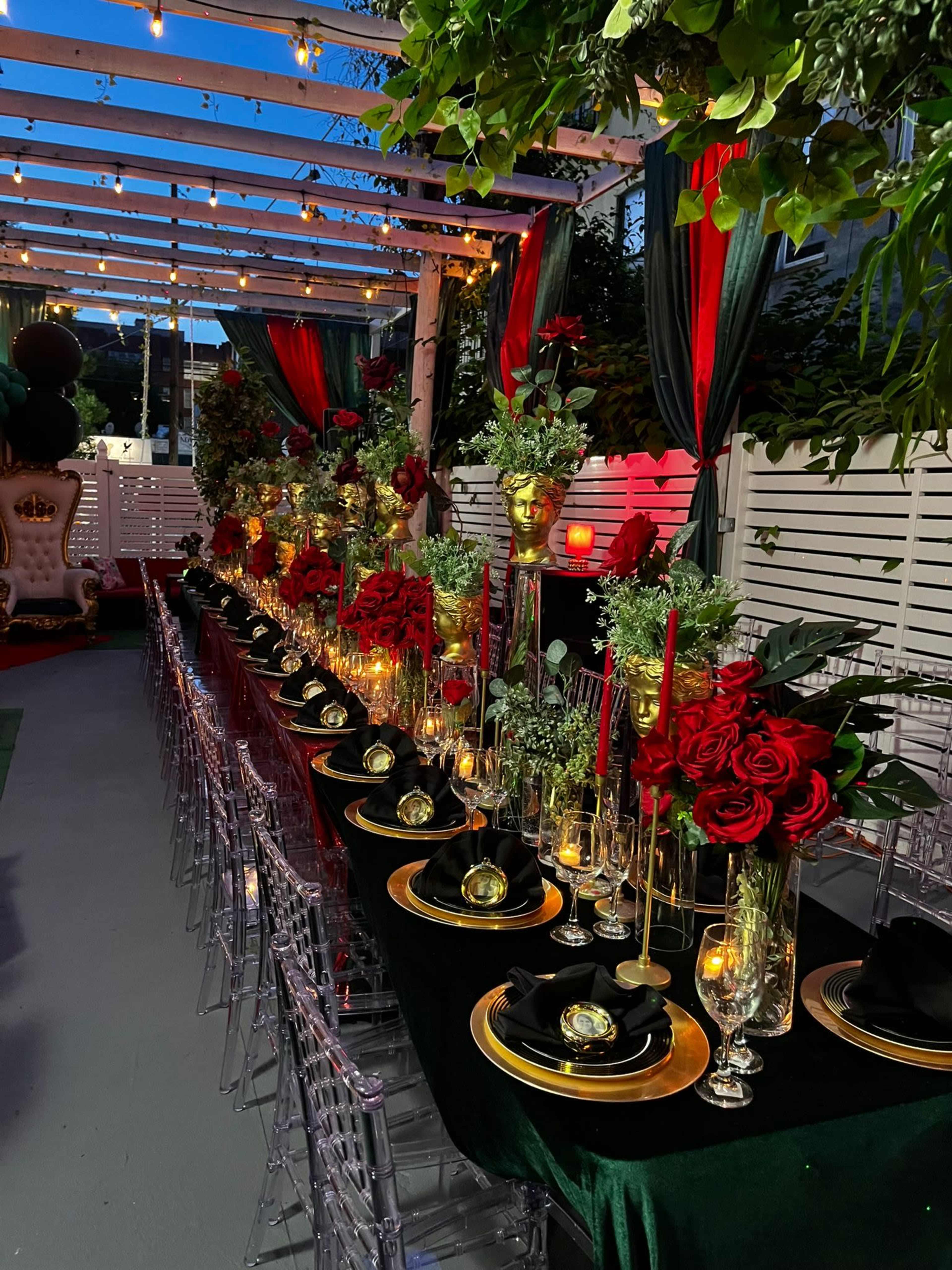 The image shows a beautifully arranged long dining table adorned with red roses, gold accents, and candles, all set under a decorated pergola with draped fabrics and string lights.