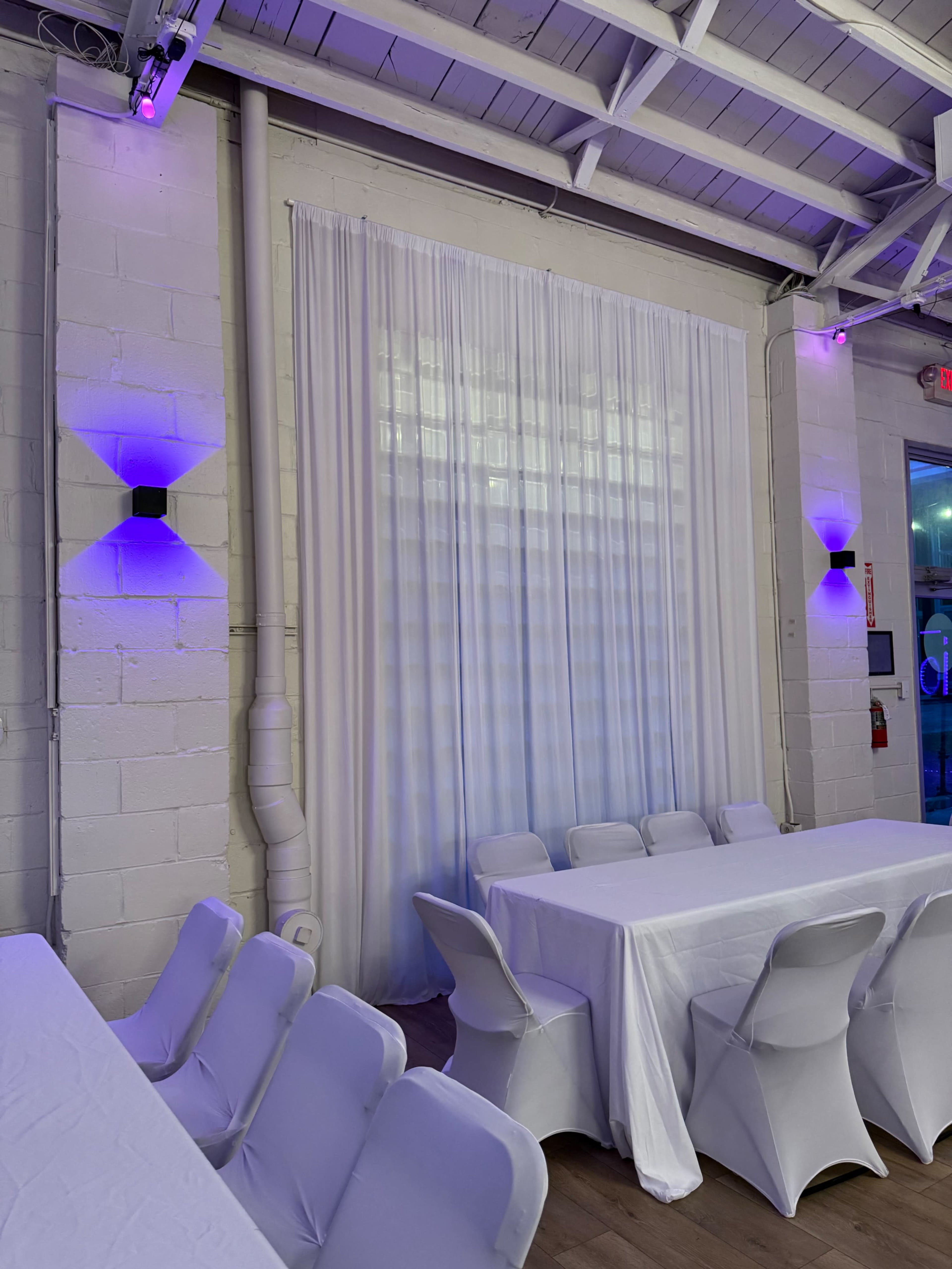 The image shows a spacious indoor venue with white tables set up and a large white curtain in the background, illuminated by purple lights on the walls.