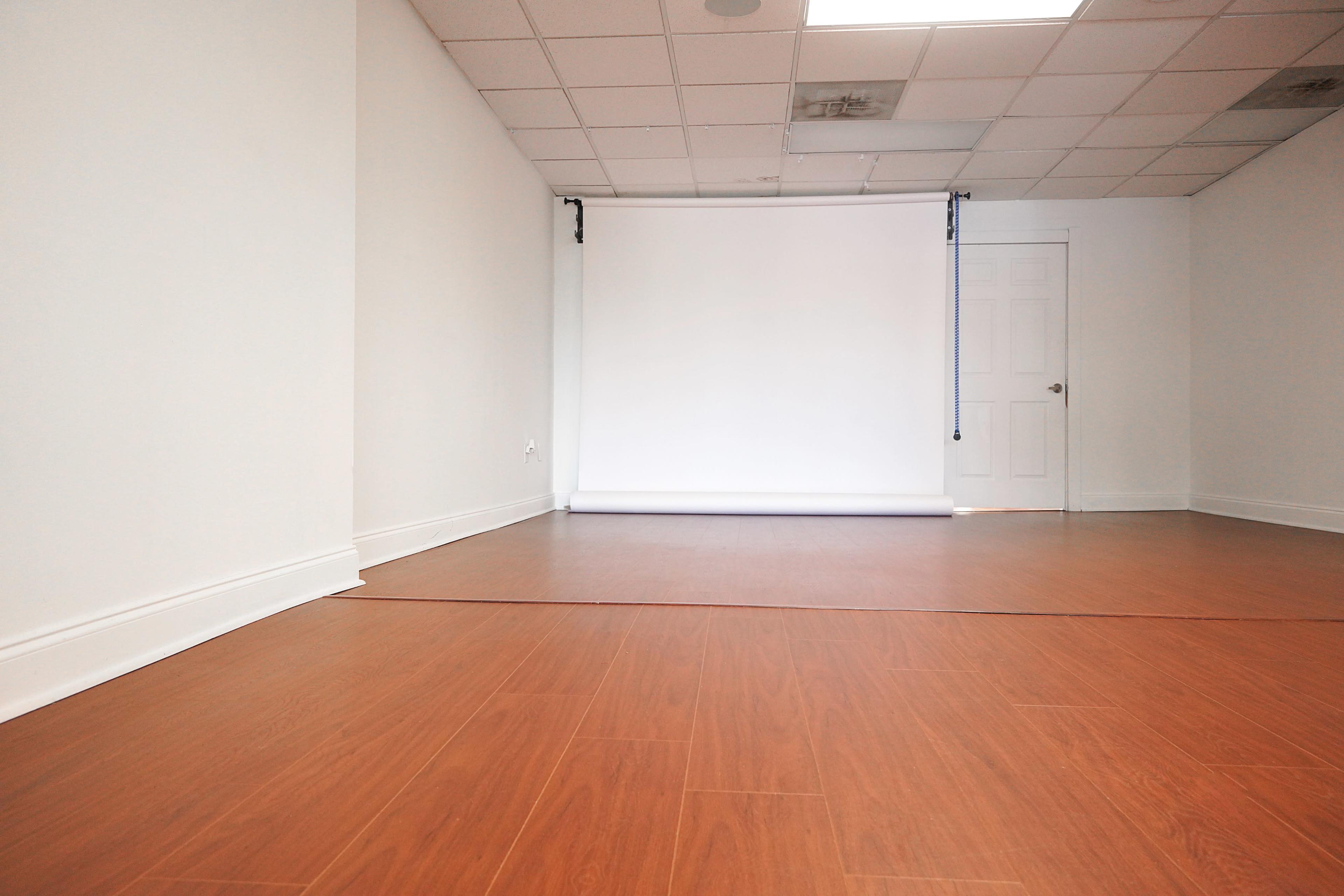 An empty room with light-colored walls and a wooden floor, featuring a white backdrop at one end and a closed door on the opposite wall.