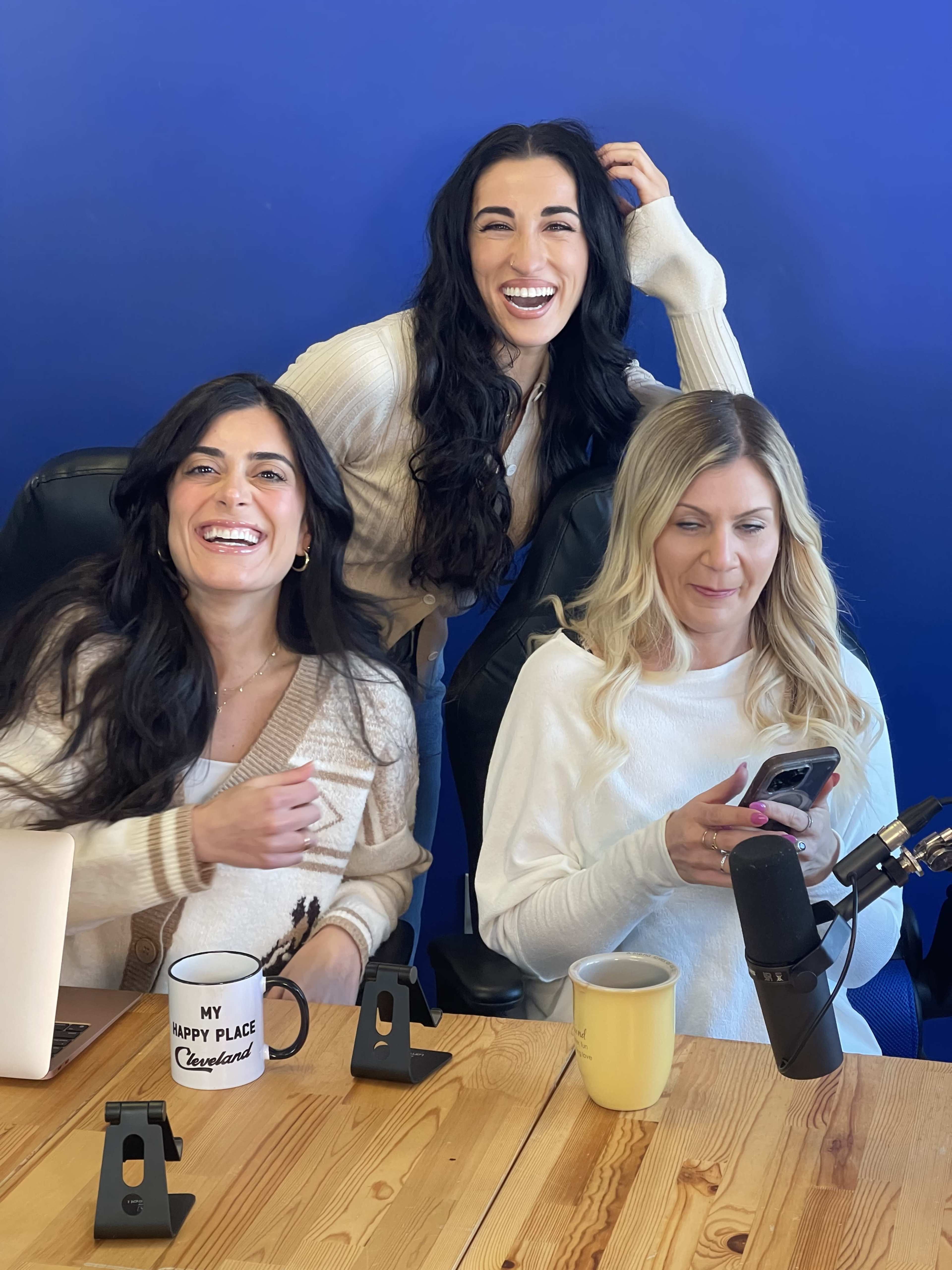 Three women sit at a wooden table, laughing together, with a blue wall backdrop and microphones in front of them.