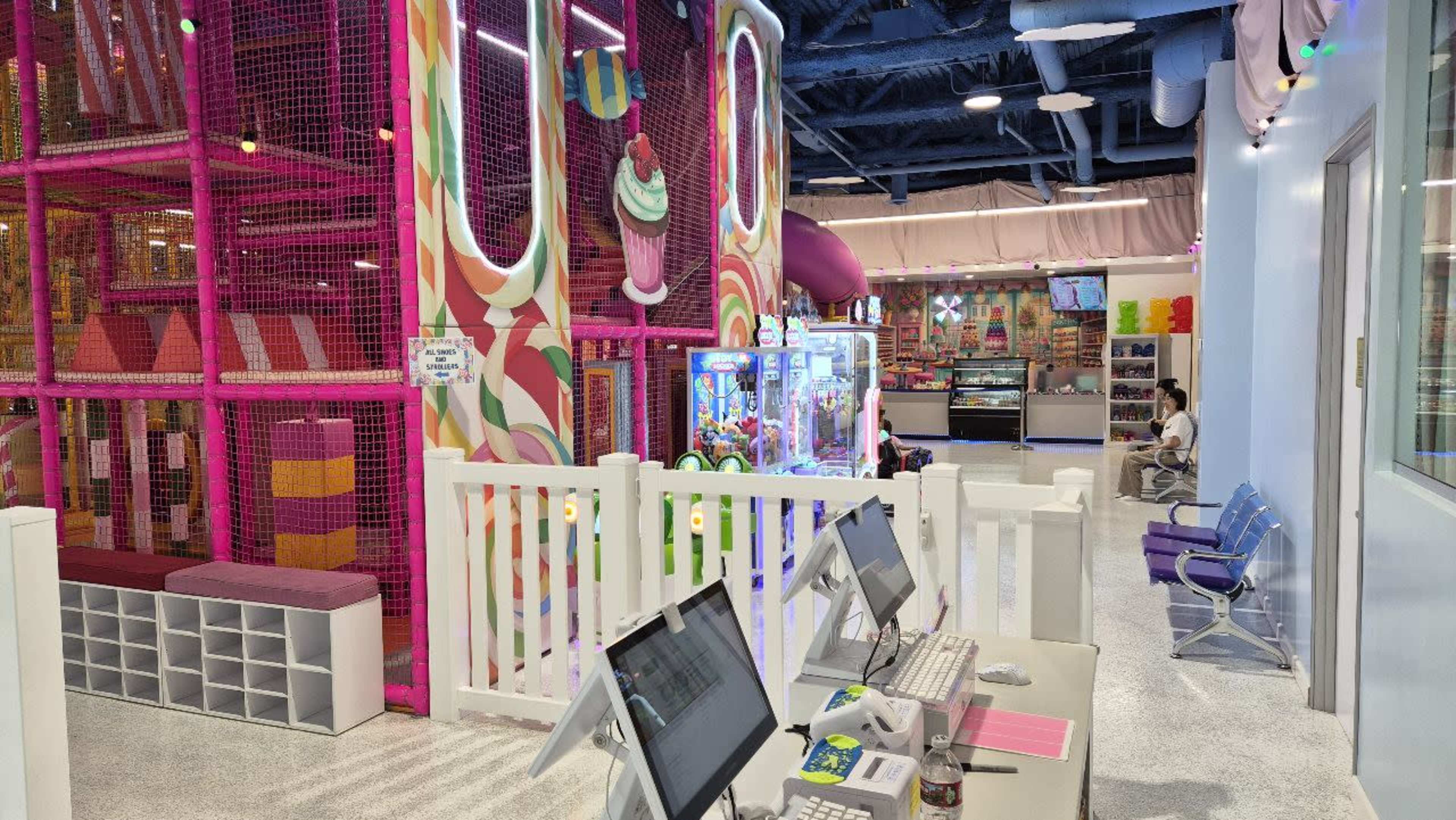 The image shows an indoor children's playground with colorful play structures, arcade machines, and a waiting area with seating.