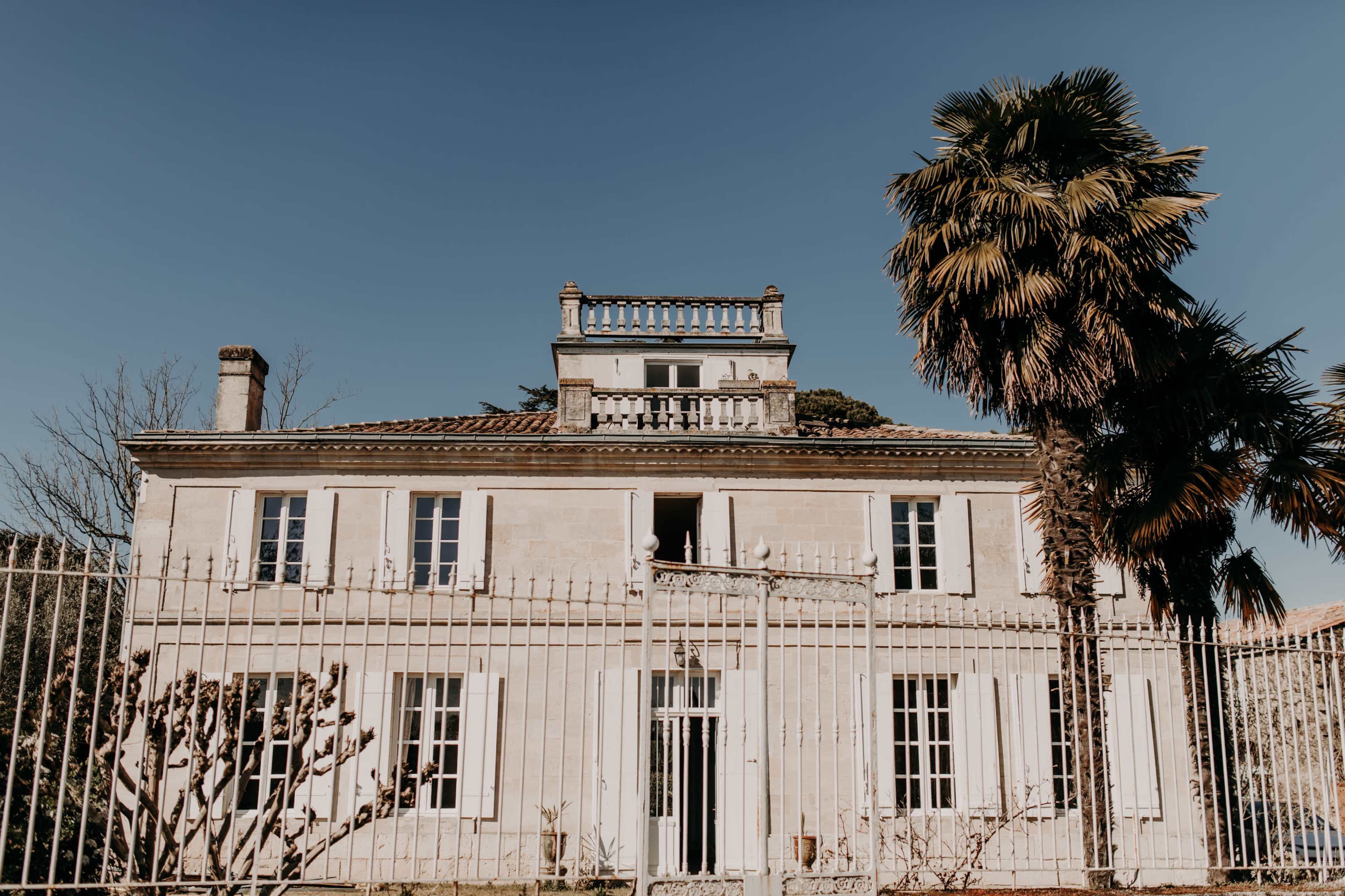 A two-story stone house with a balcony and palm trees in front, enclosed by a white metal fence.