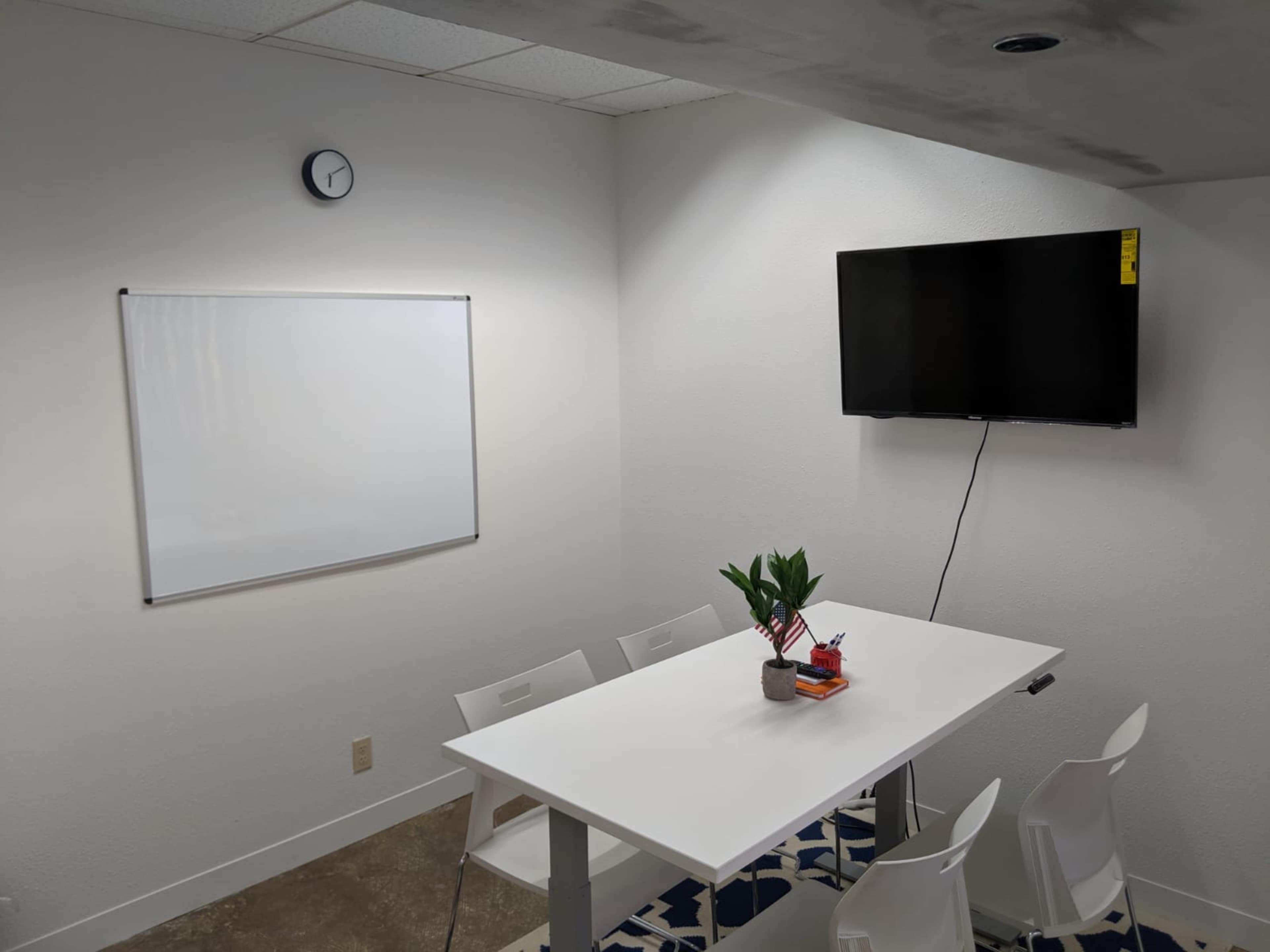 A meeting room features a whiteboard, a clock, a television on the wall, and a table surrounded by chairs.