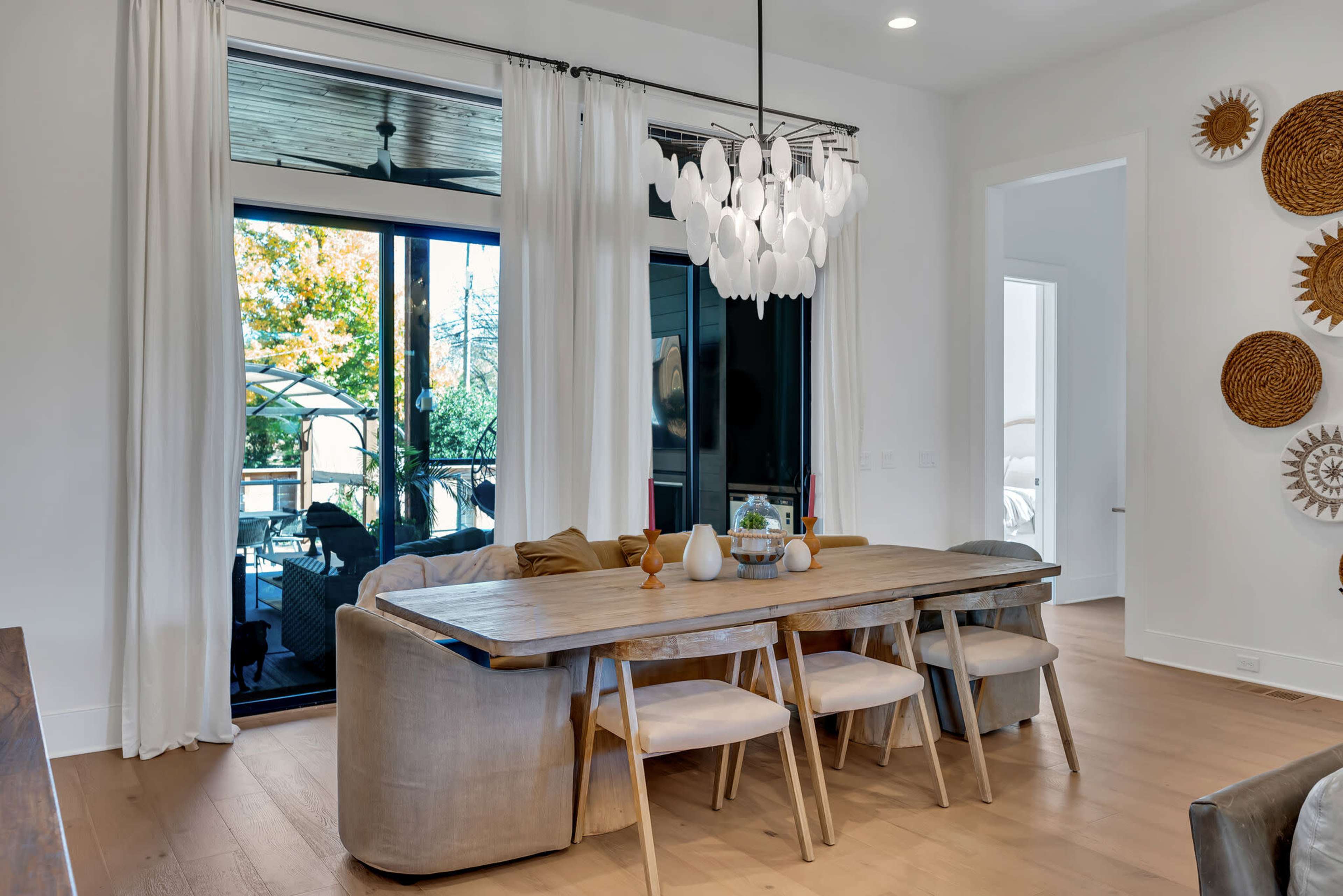 A wooden dining table with three chairs is positioned in a brightly lit room adorned with white curtains and decorative wall hangings.