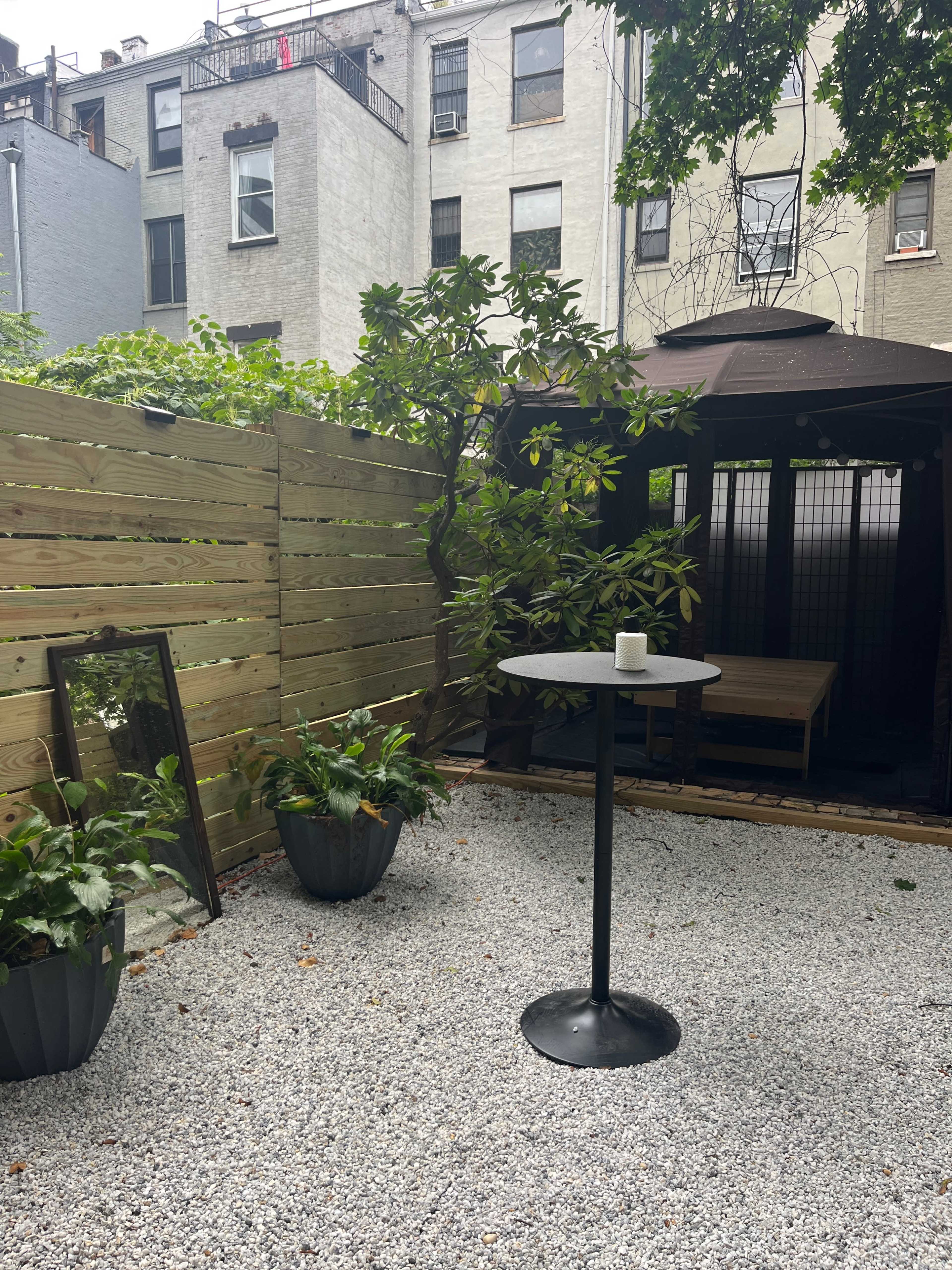 A small outdoor garden area featuring a gravel surface, a black standing table with a candle, potted plants, and a gazebo in the background amidst wooden fencing.