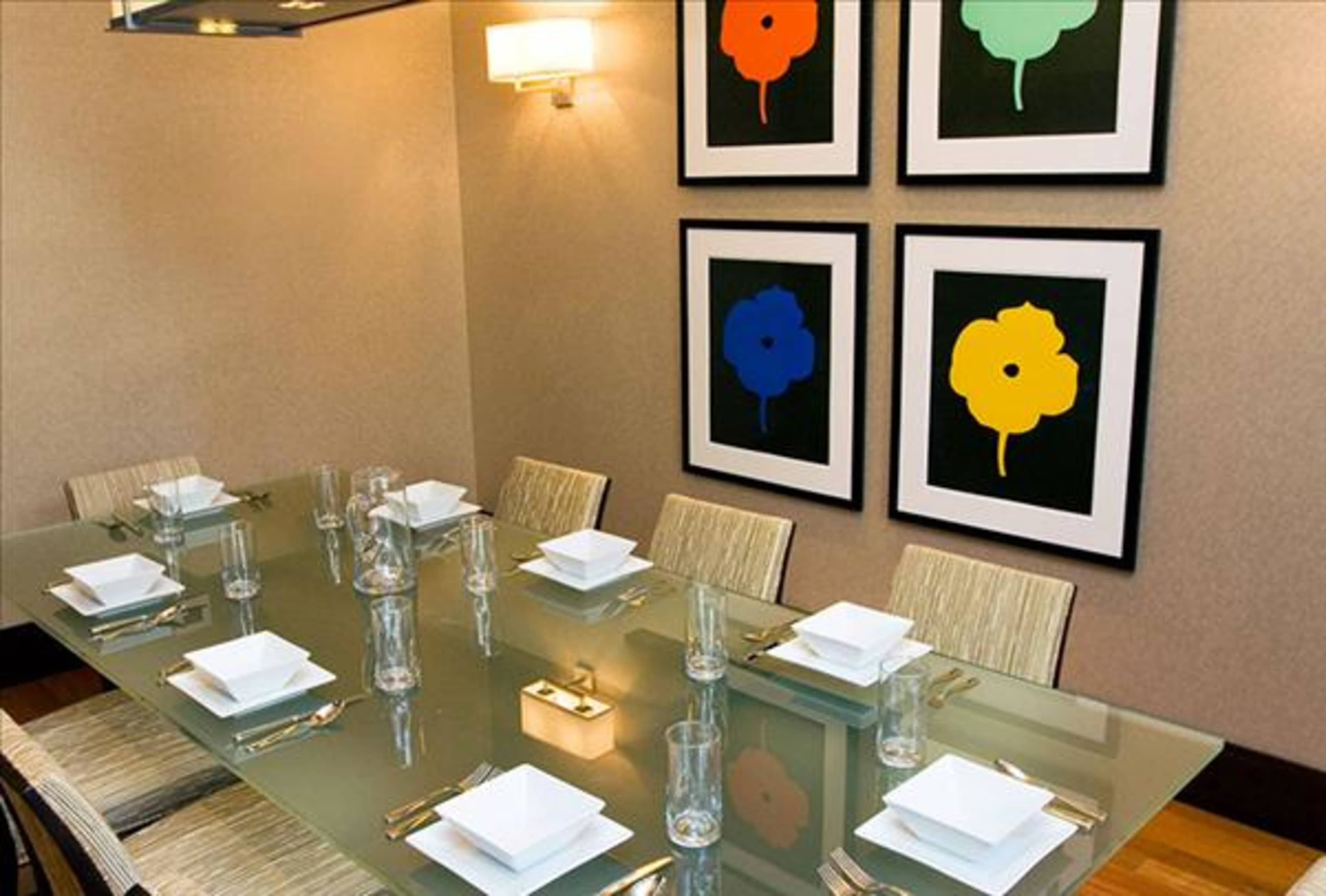 A modern dining room features a glass table set with white dishes and glasses, surrounded by framed colorful floral prints on the walls.