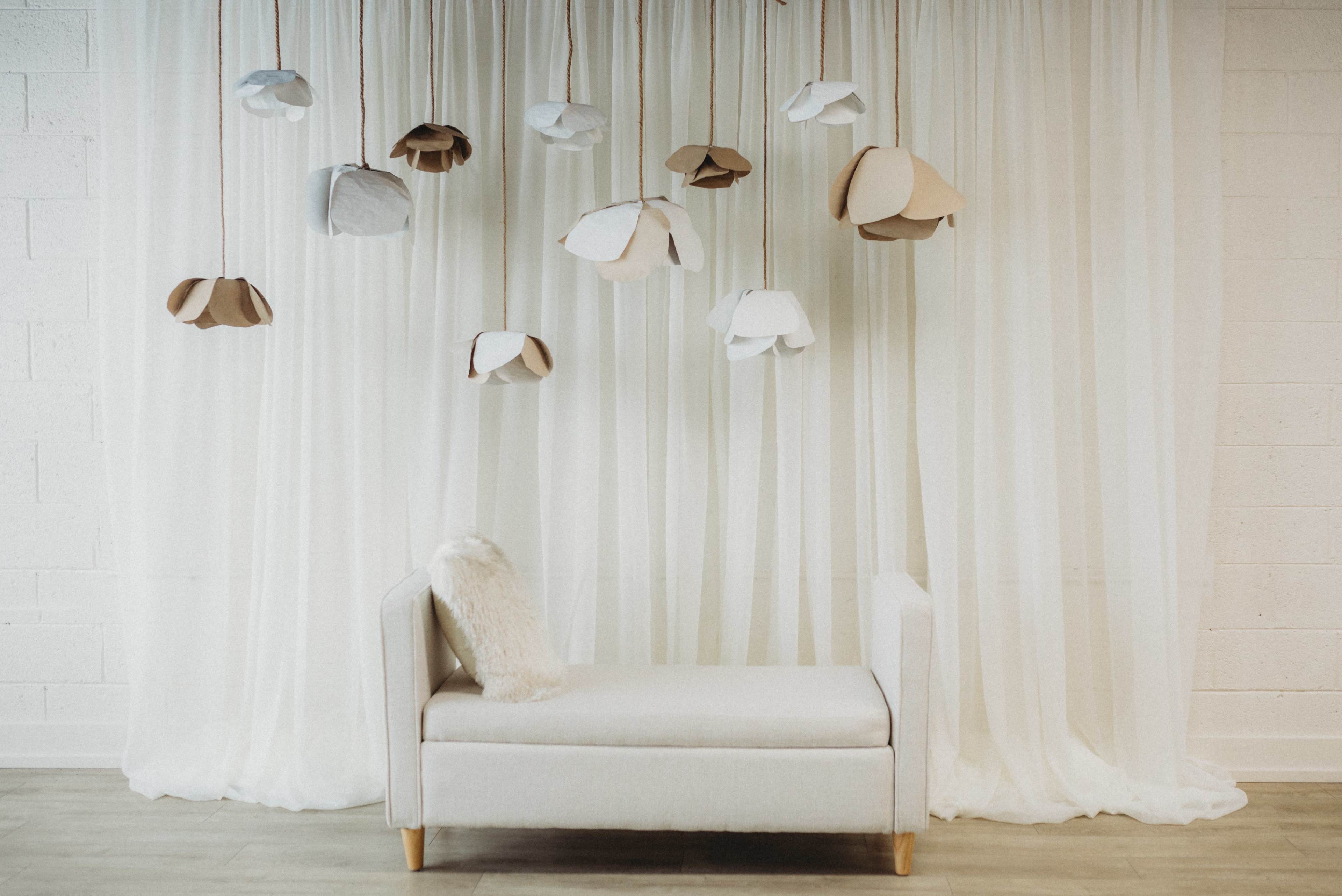 The image features a minimalistic room with a cream-colored chaise lounge beneath a backdrop of flowing white curtains and hanging paper flower decorations.