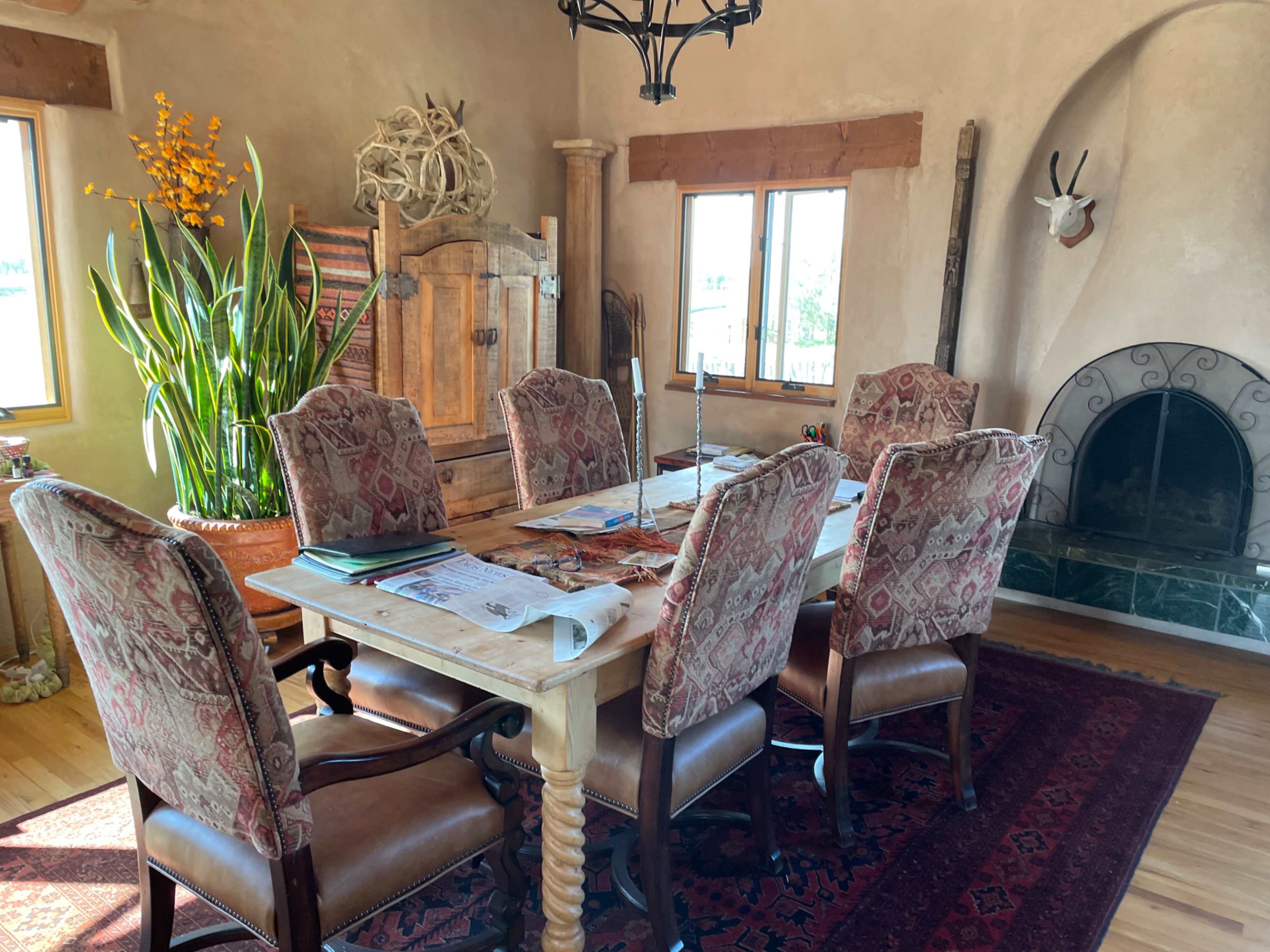 A wooden dining table with six upholstered chairs is set in a room featuring large windows, a green marble fireplace, and decorative plants.