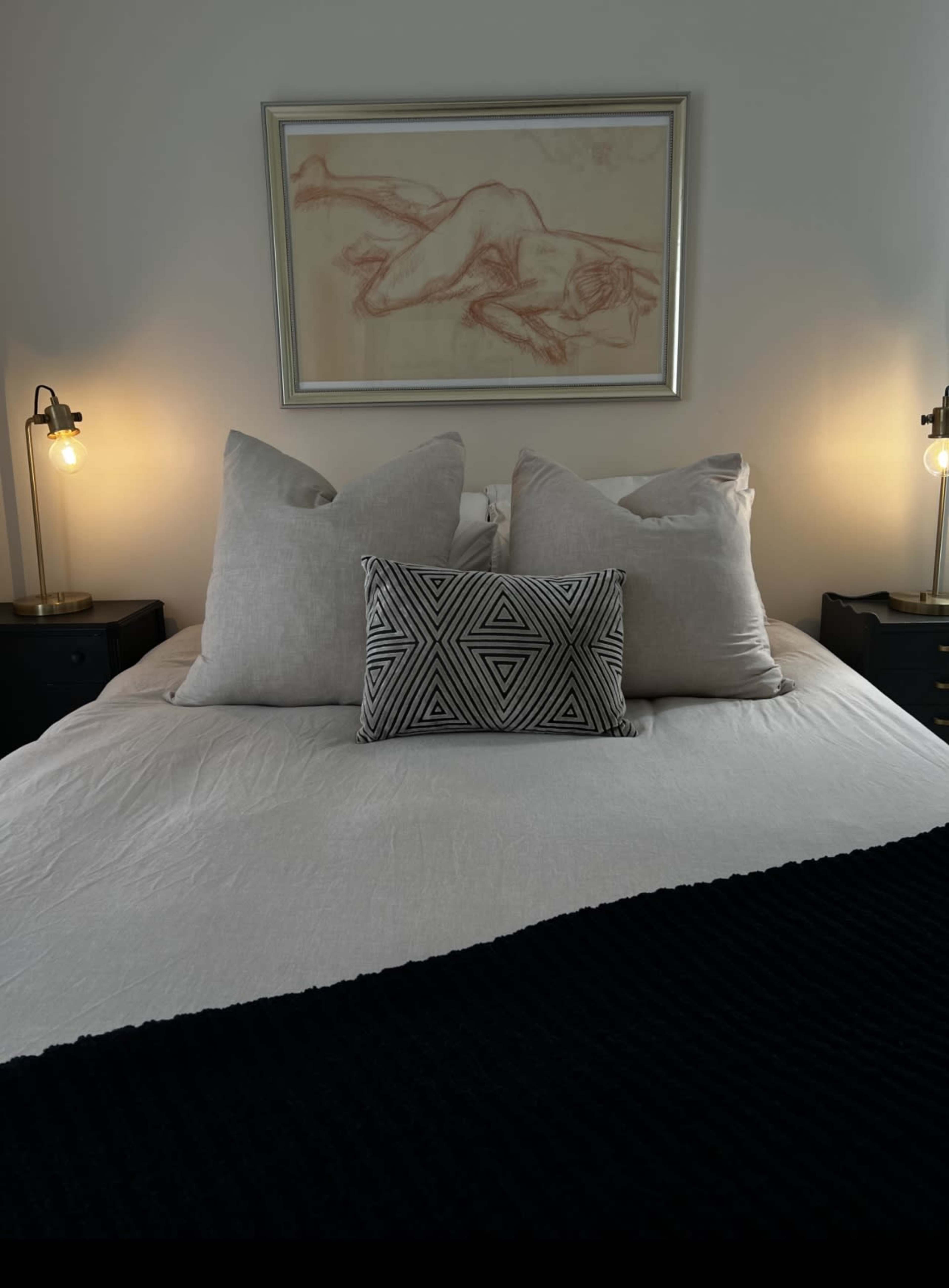 The image shows a neatly made bed with several decorative pillows, a black throw, and framed artwork of a reclining figure on the wall above.