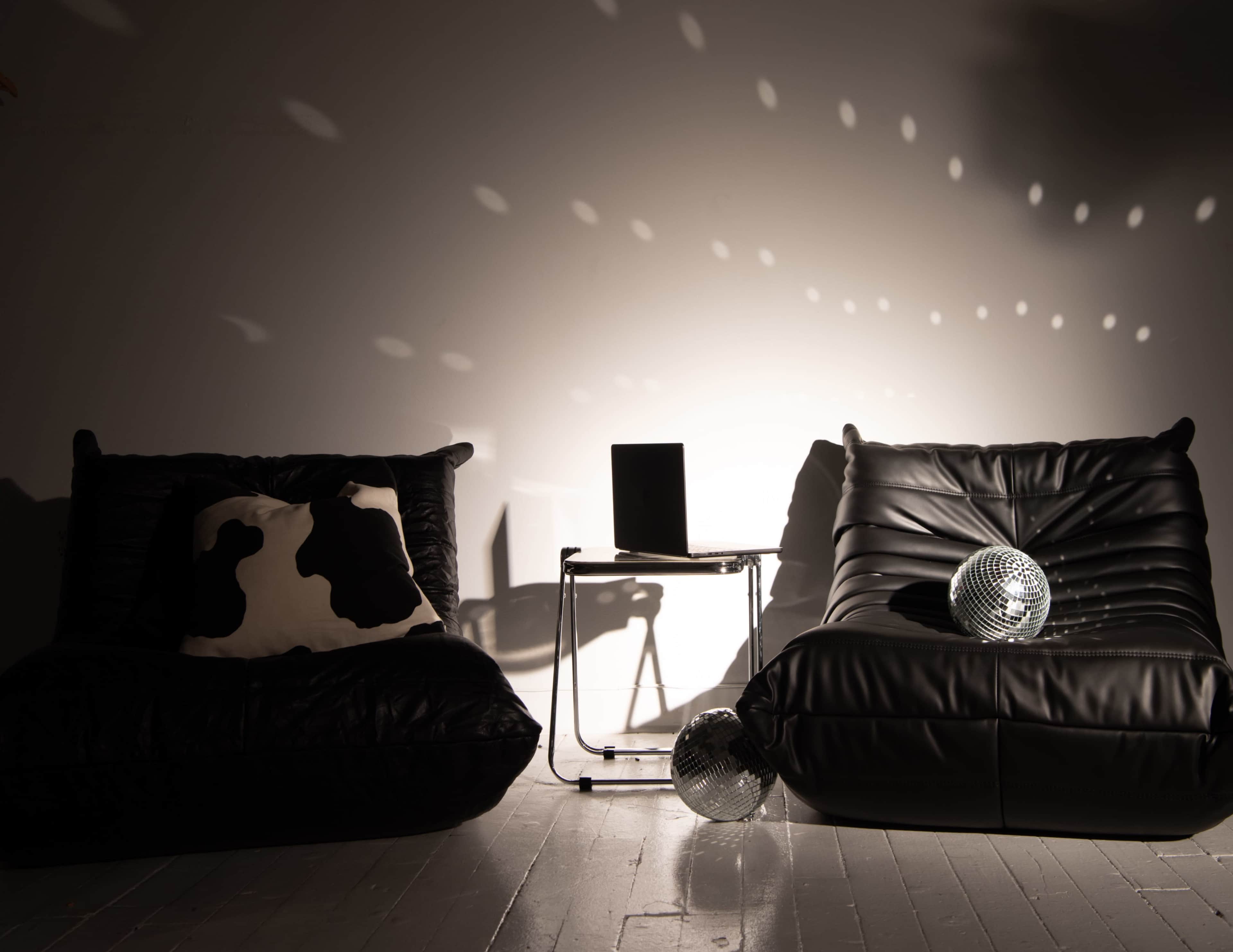 The image features two black lounge chairs with a cow-print pillow, a small table, and two mirrored disco balls on a light-colored floor, illuminated by spotlights that create circular reflections on the wall.