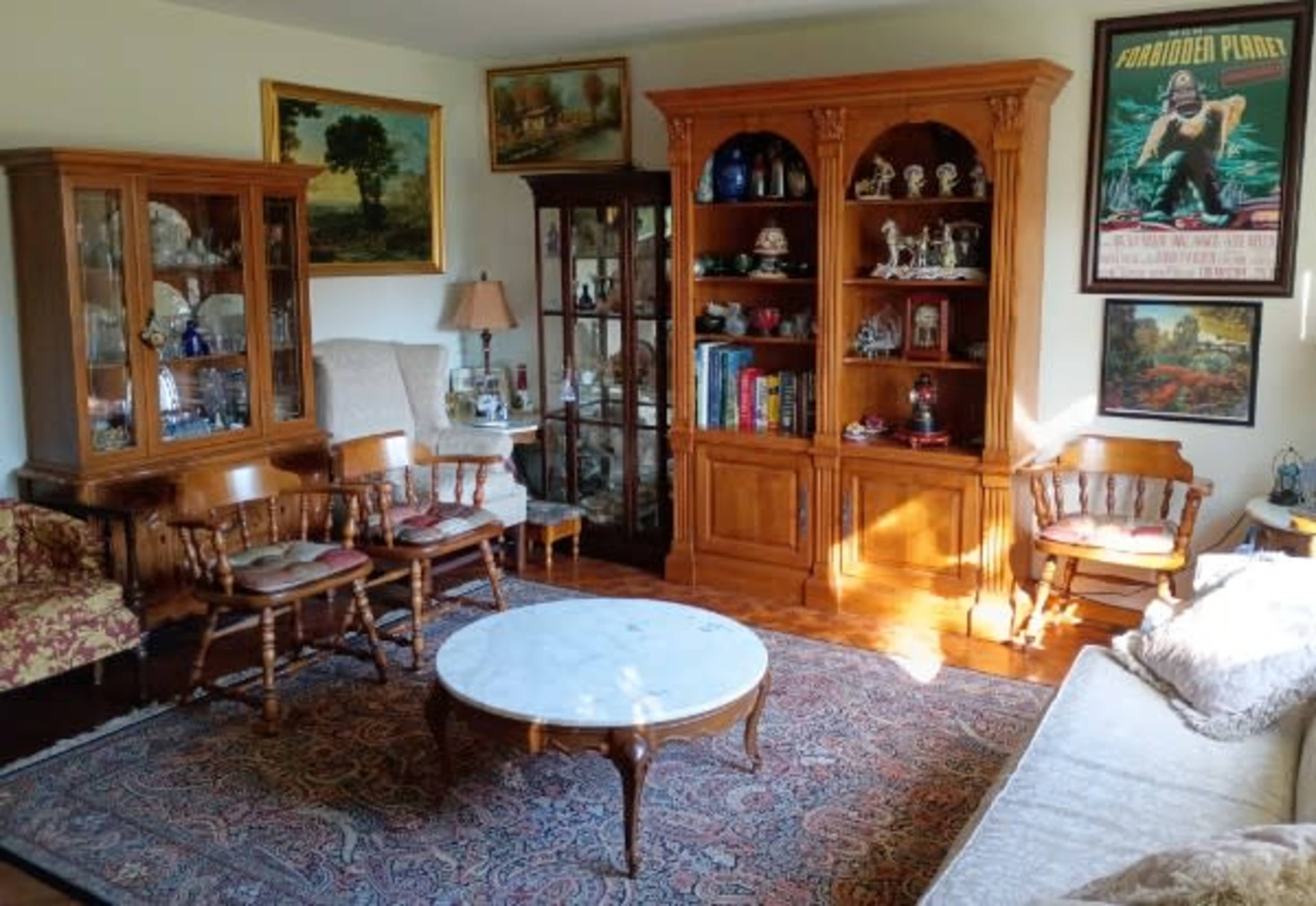 The image shows a cozy living room featuring wooden furniture, a large area rug, and various decorative items displayed on shelves and in glass cabinets.