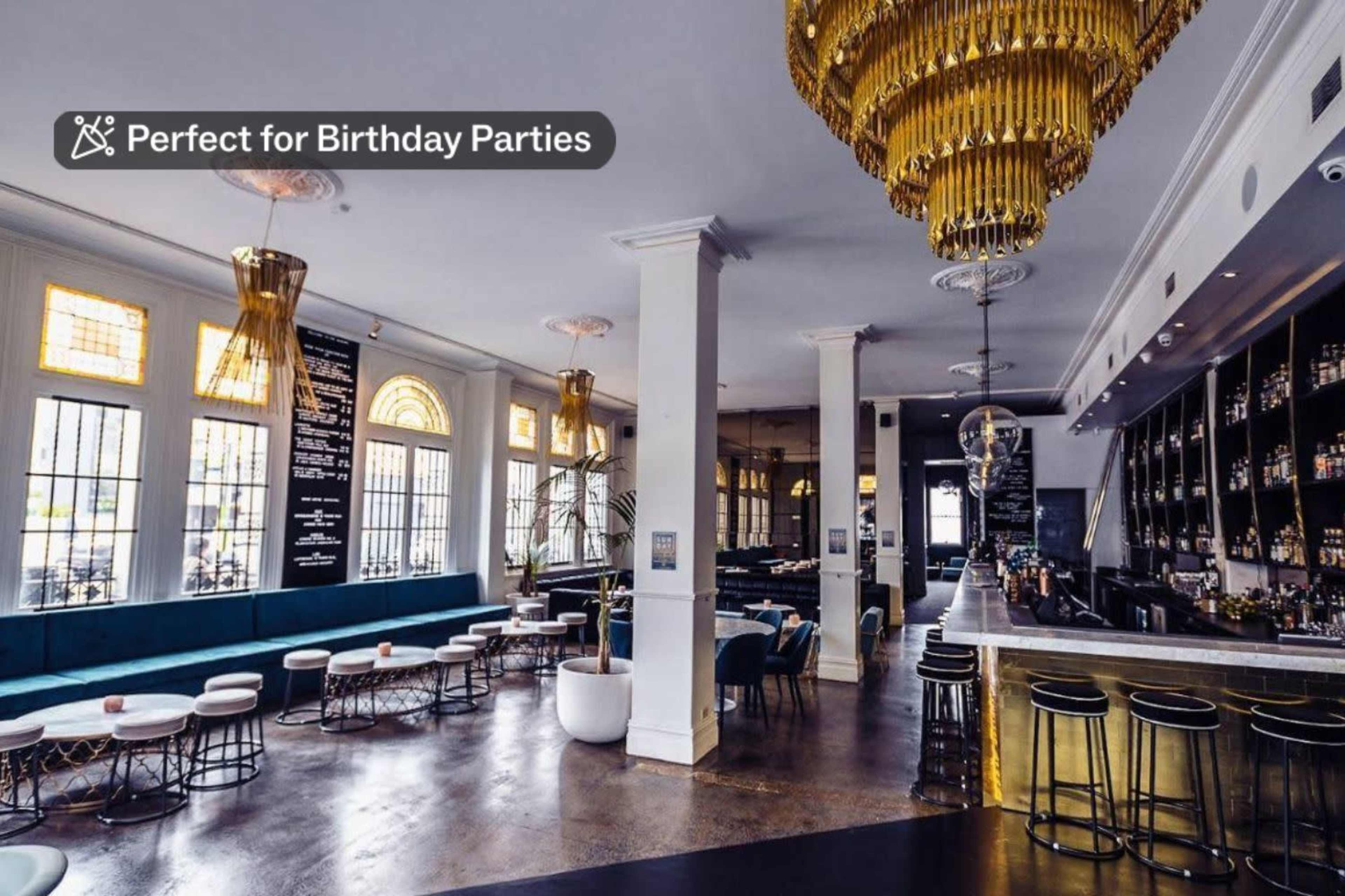 A stylish bar interior with golden chandeliers, a sleek bar area, and blue lounge seating arranged around small tables.