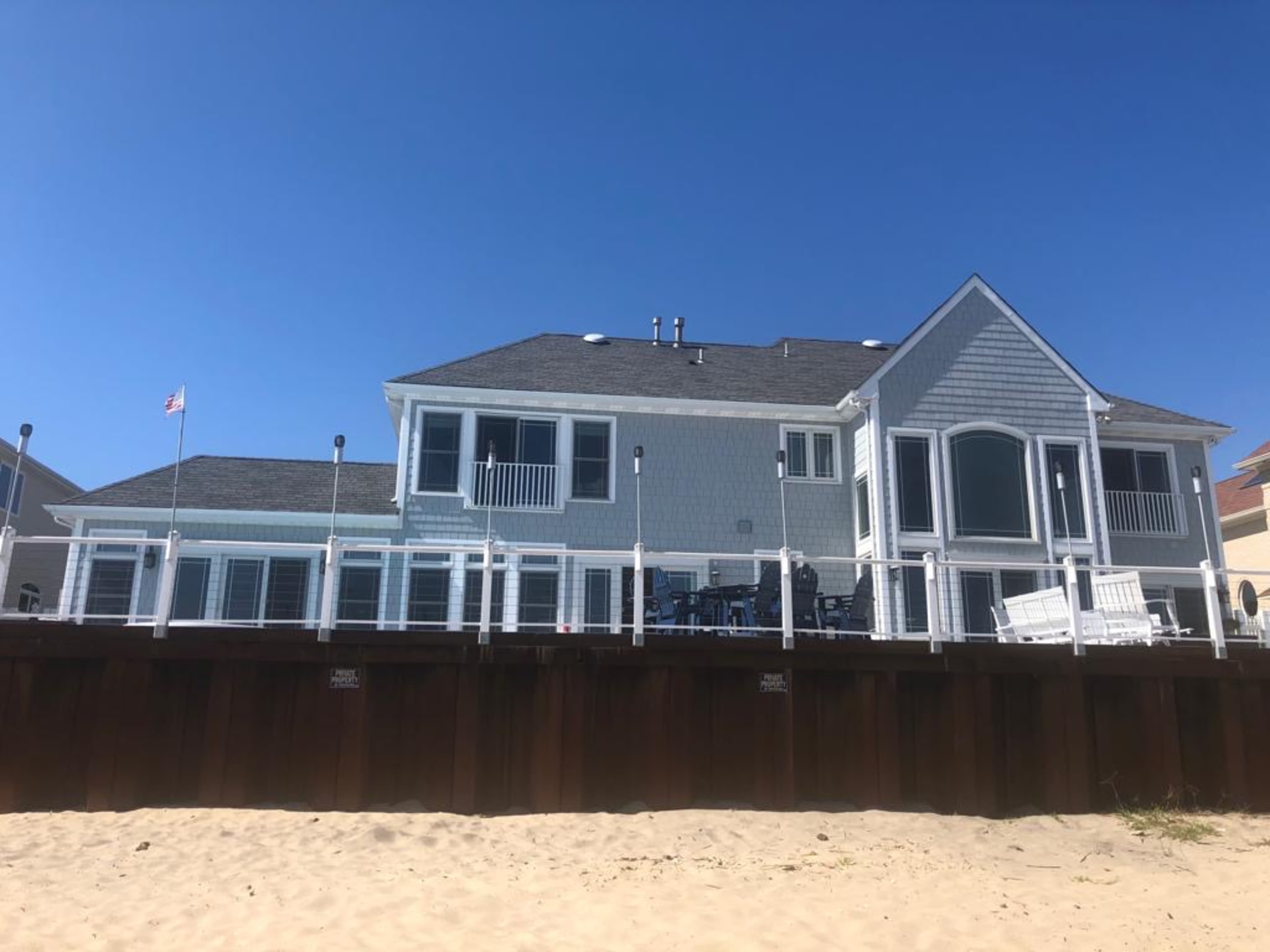 A beach house with a gray exterior and a wooden deck overlooks a sandy shore under a clear blue sky.