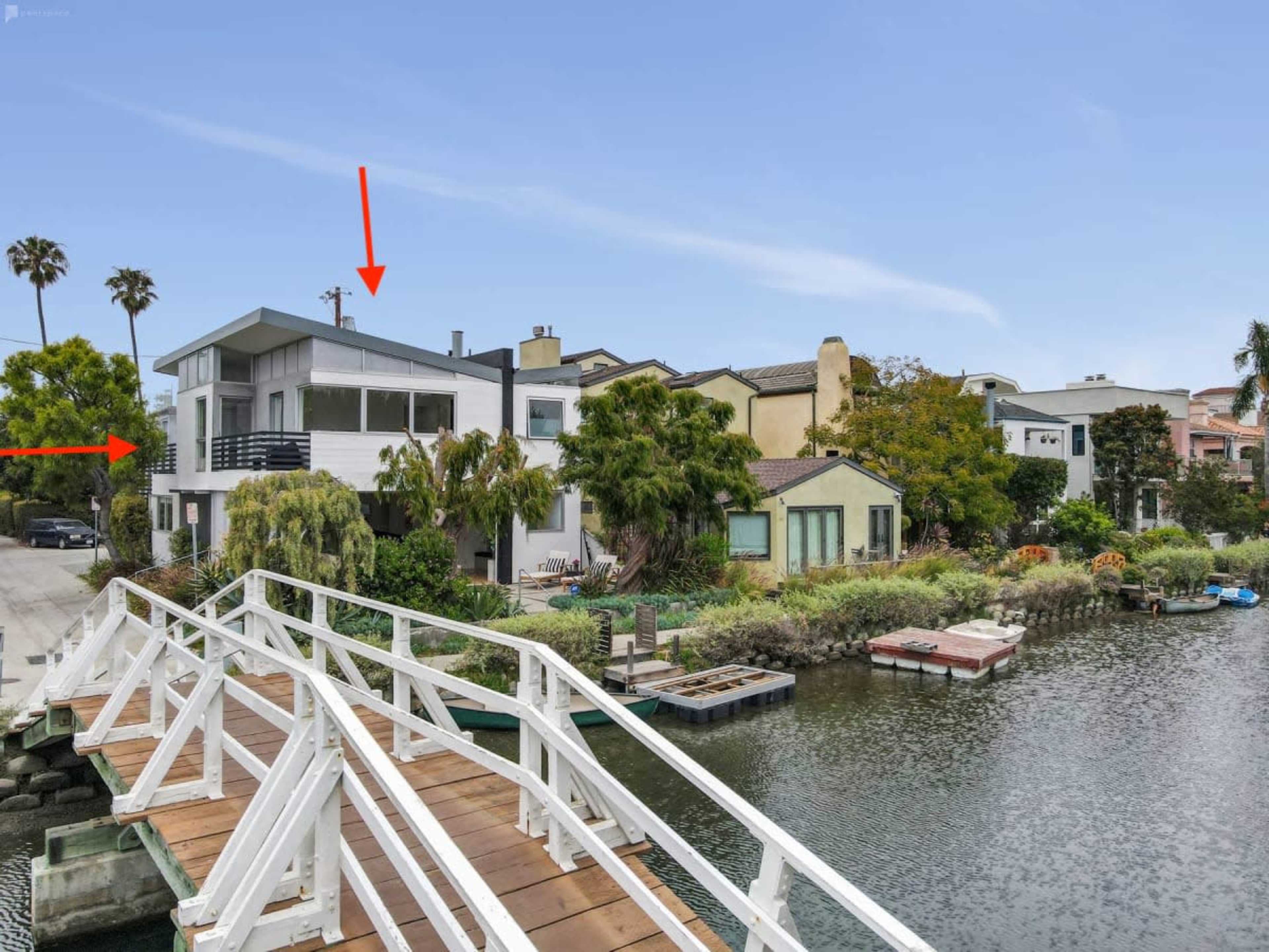 A modern house with large windows is situated by a canal, with a wooden walkway leading to it.
