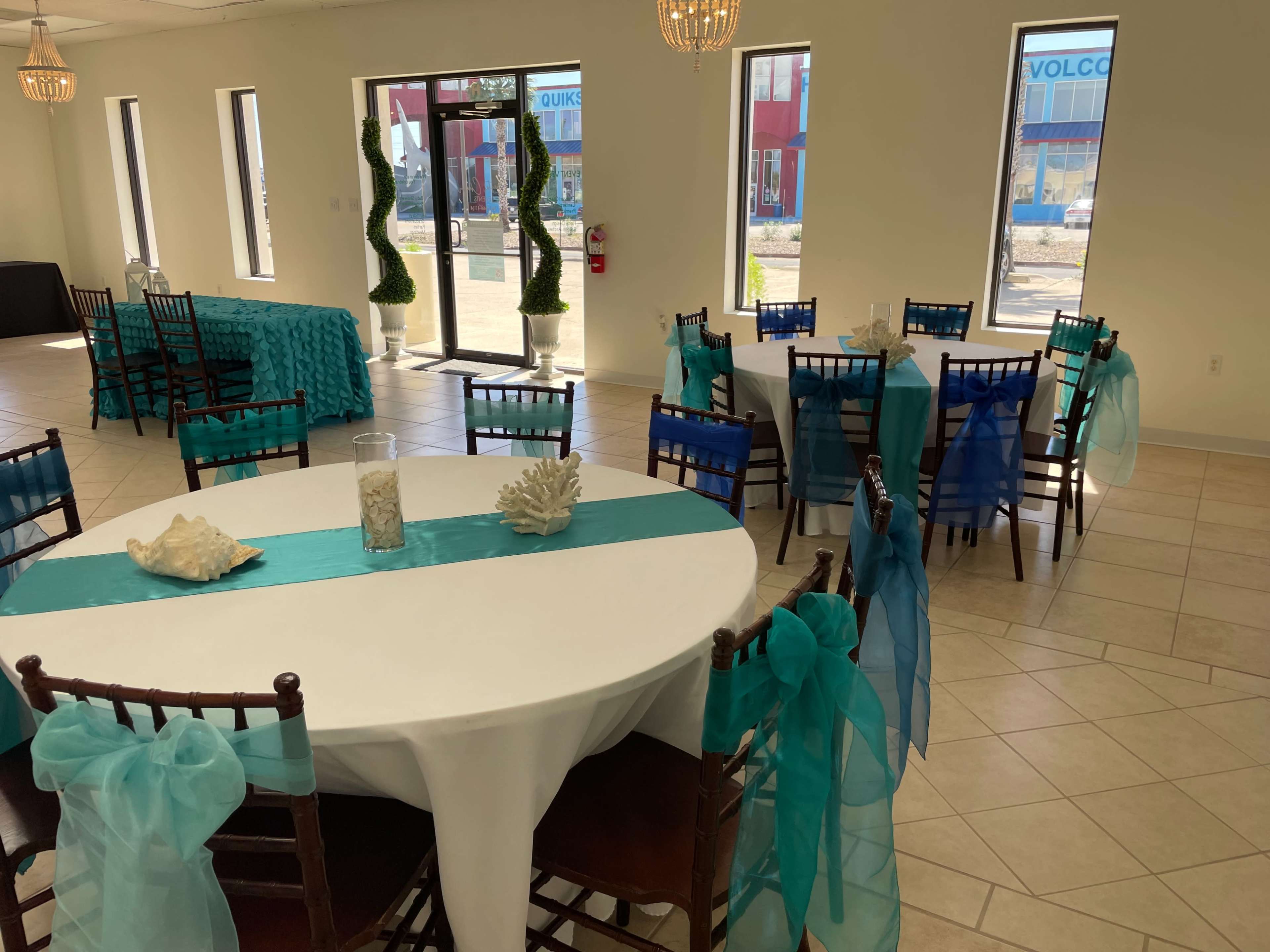 The interior of an event space features round tables with white tablecloths and teal accents, accompanied by chairs adorned with teal sashes, with large windows allowing natural light in.