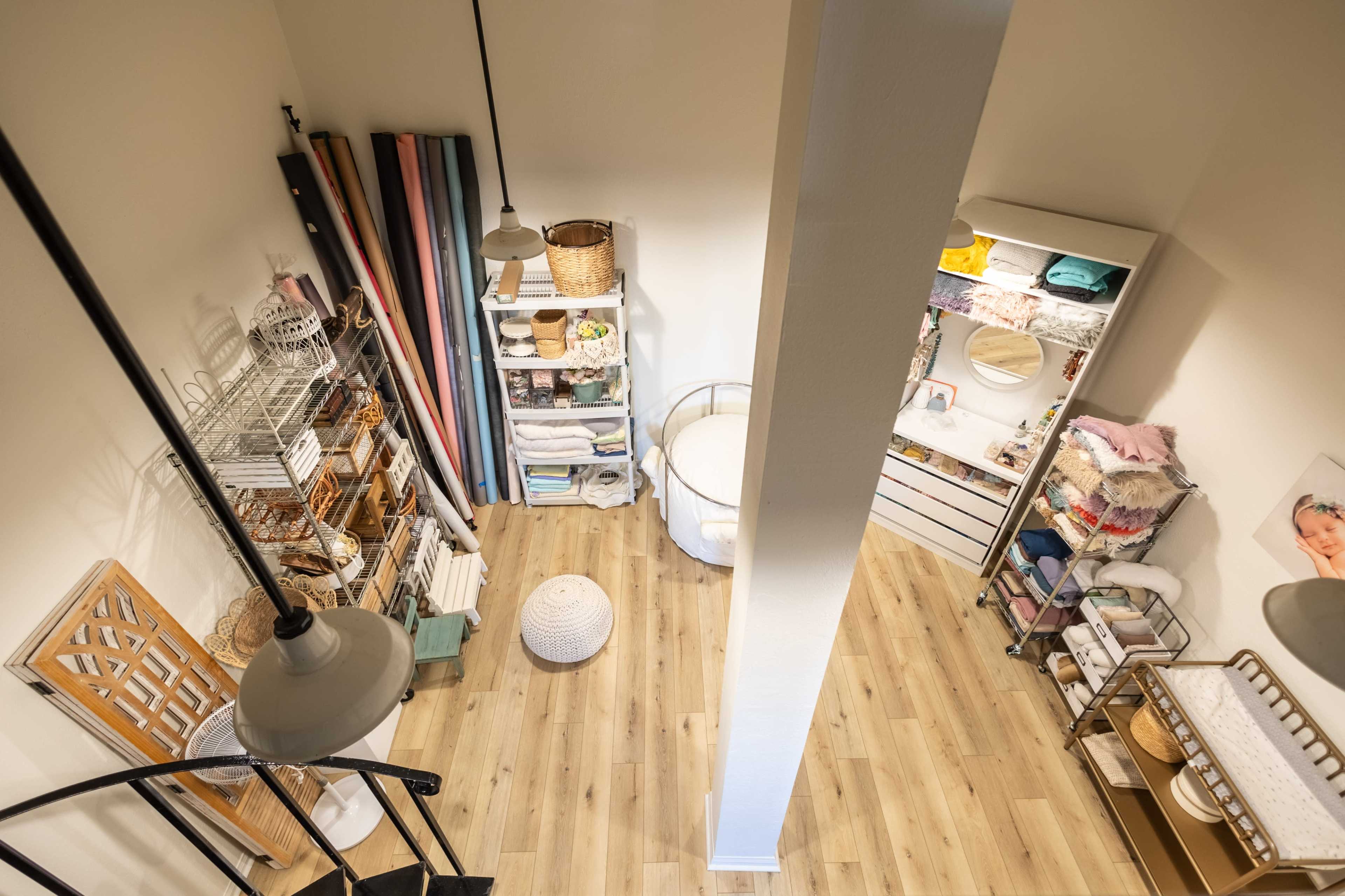 The image shows a small, organized storage room with shelves containing baskets, linens, and a round pouf on the wooden floor.