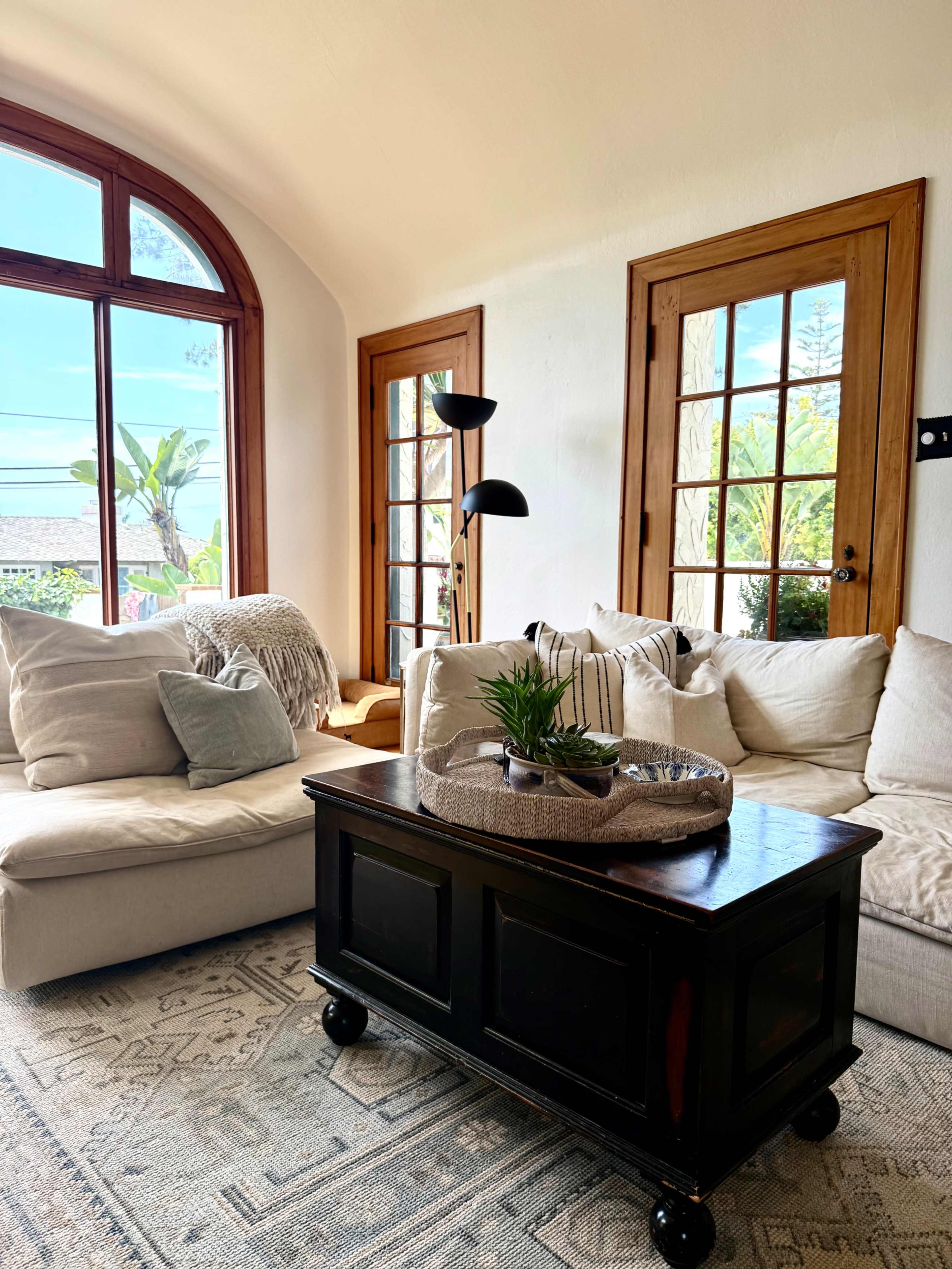 A cozy living room features a light-colored sectional sofa, a dark wood coffee table with a decorative tray, and large windows with a view of greenery.