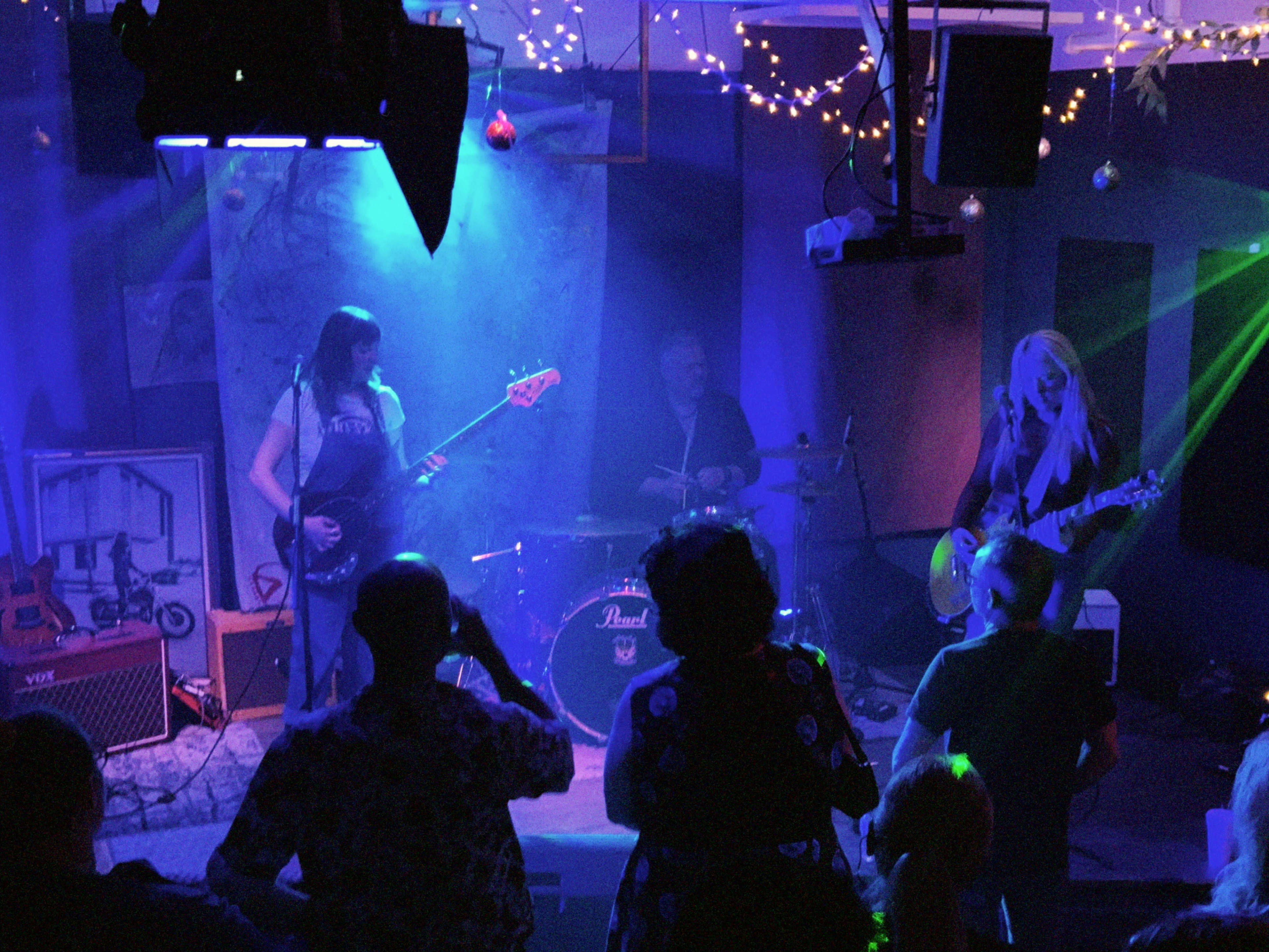 A band performs on stage in a dimly lit venue with colorful lighting, while an audience watches attentively.