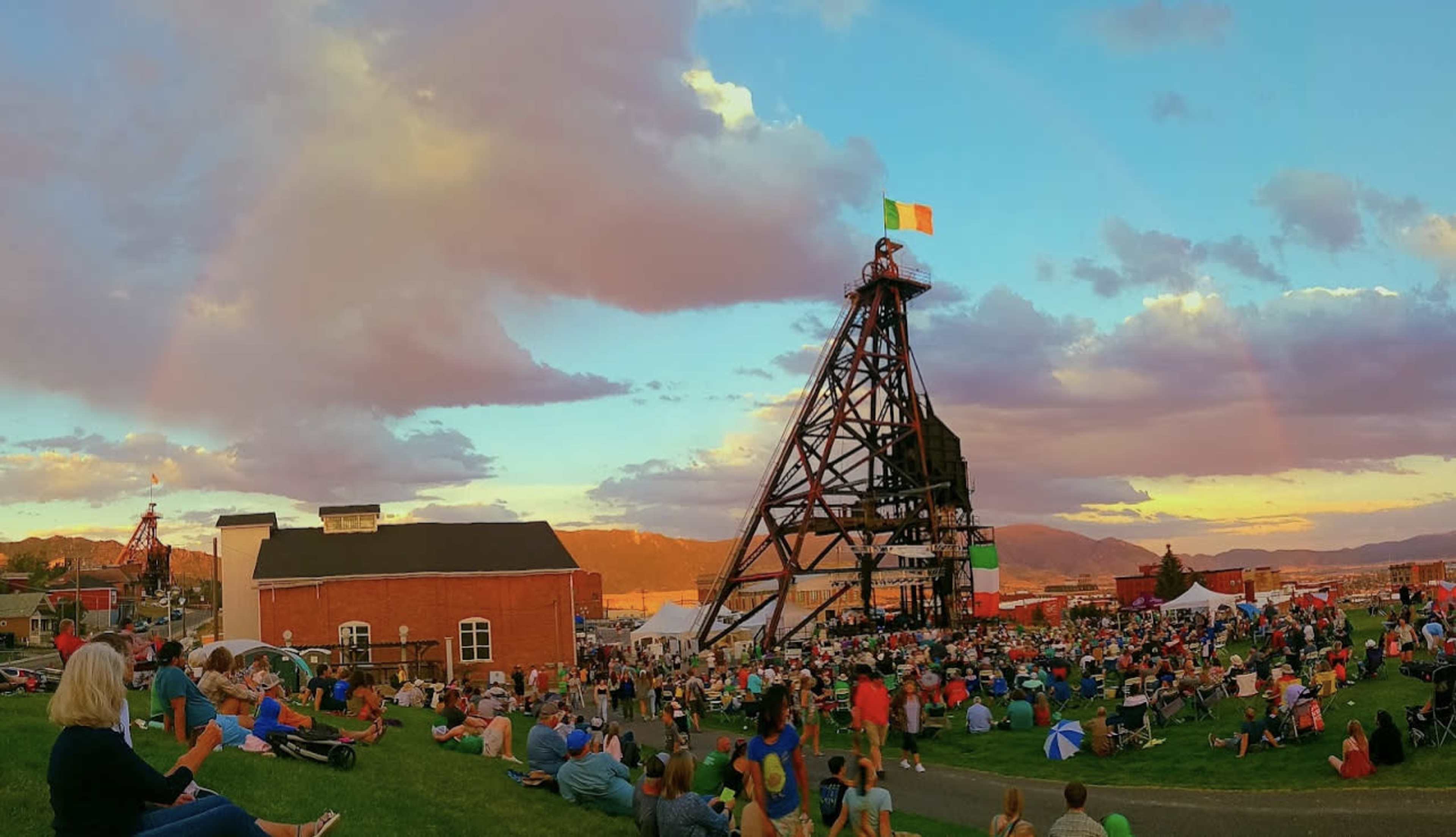 A large crowd gathers on a grassy hillside in front of a historic mining structure during an outdoor event, with colorful flags and a rainbow in the sky.