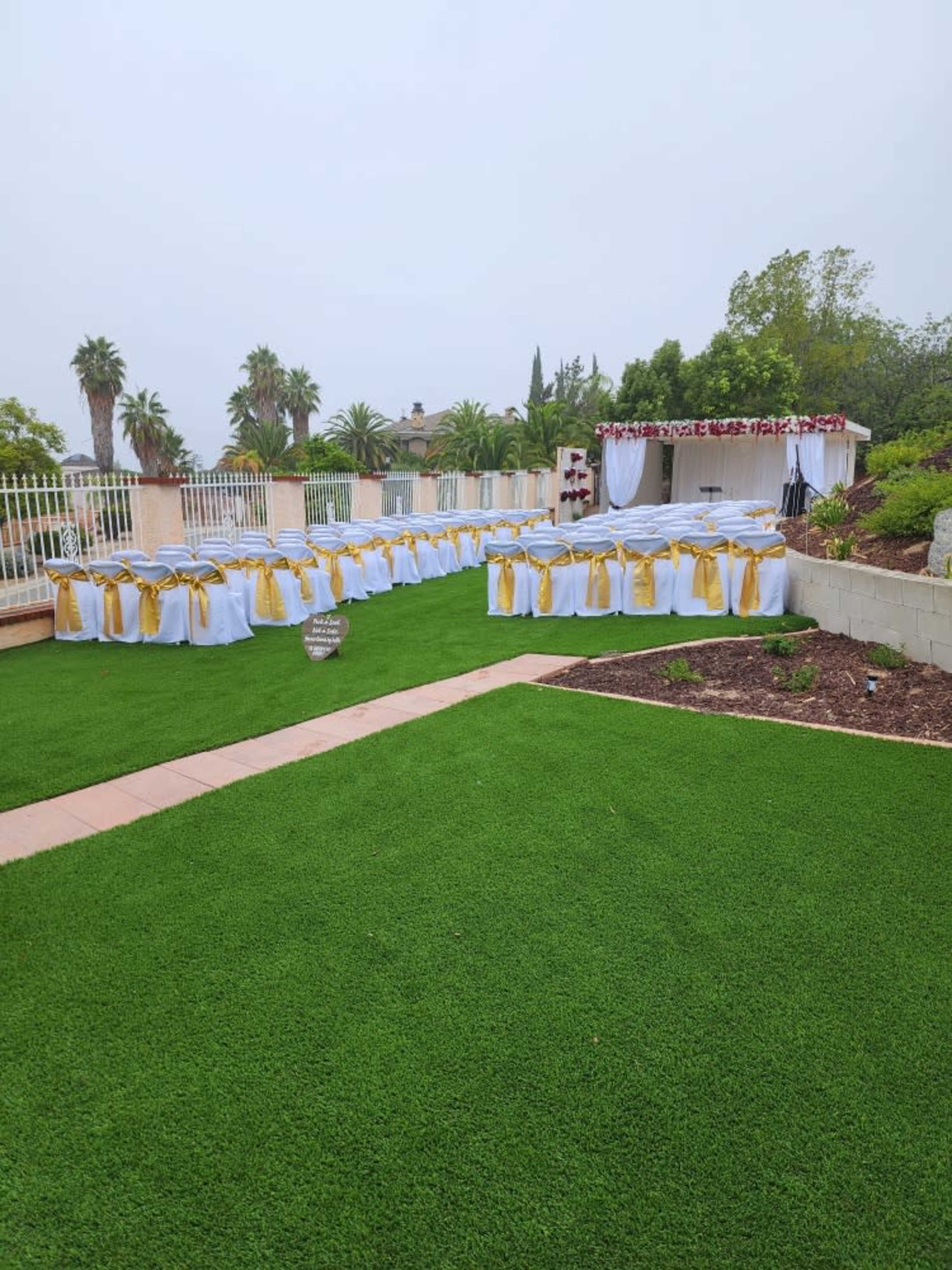 A beautifully arranged outdoor event space features tables dressed in white tablecloths with gold sashes, set up on vibrant green grass.