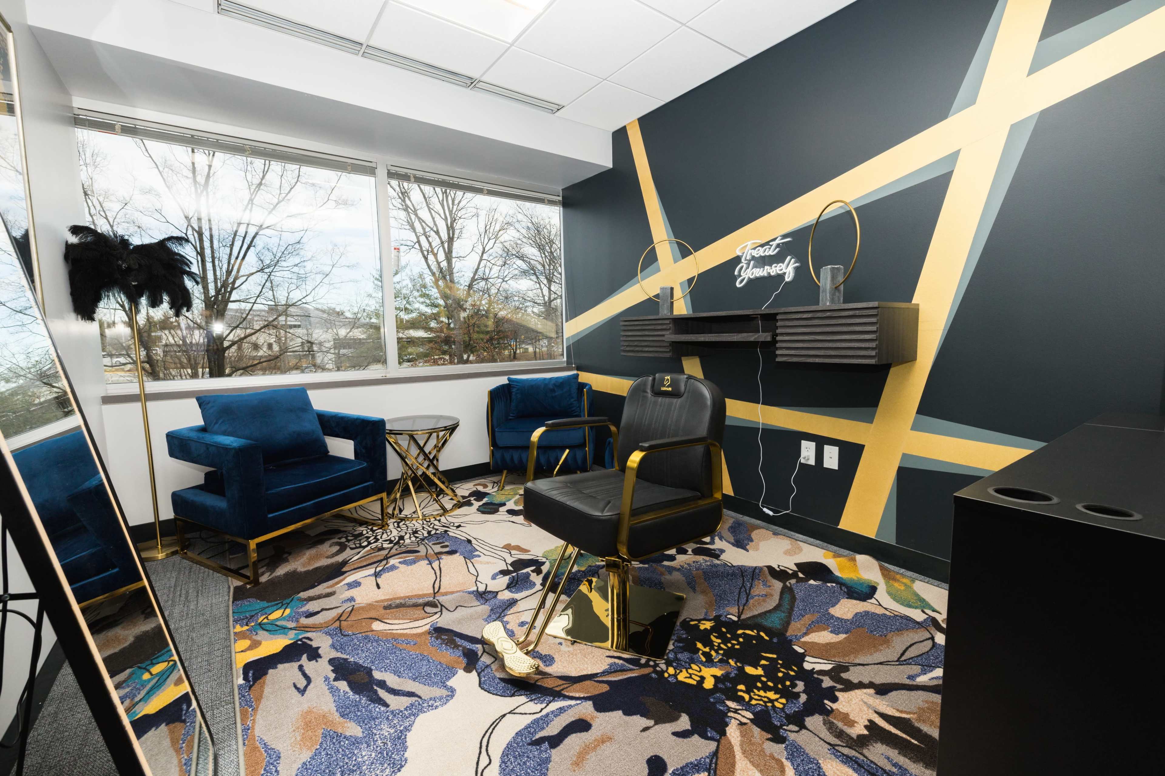 A modern office space featuring blue velvet chairs, a gold-accented chair, and a decorative rug with floral patterns, complemented by a large window and a stylish wall design.