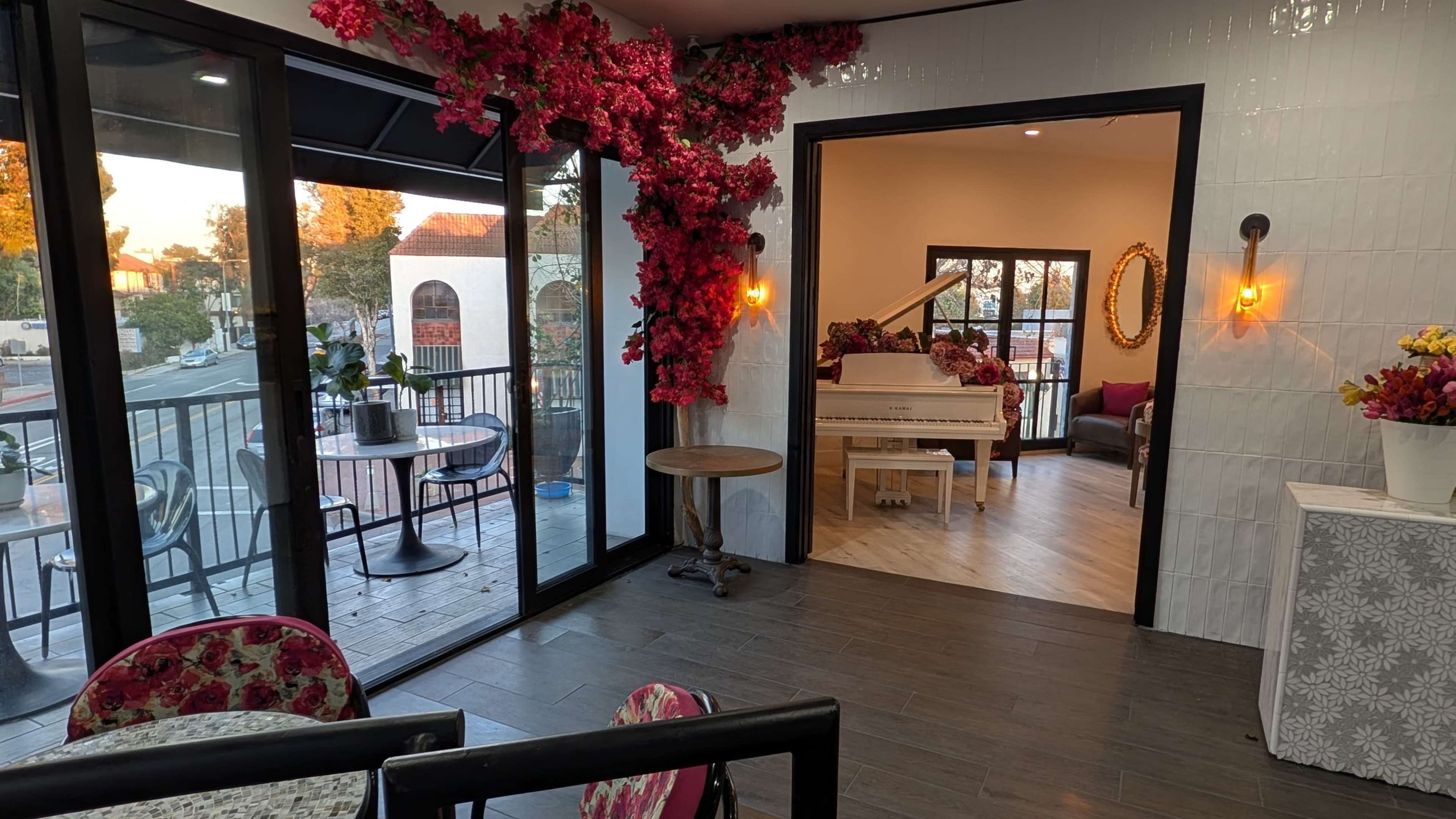 The scene shows a cozy cafe space with large glass windows, a decorative floral arrangement on the wall, and an adjoining room featuring a white piano and seating area.